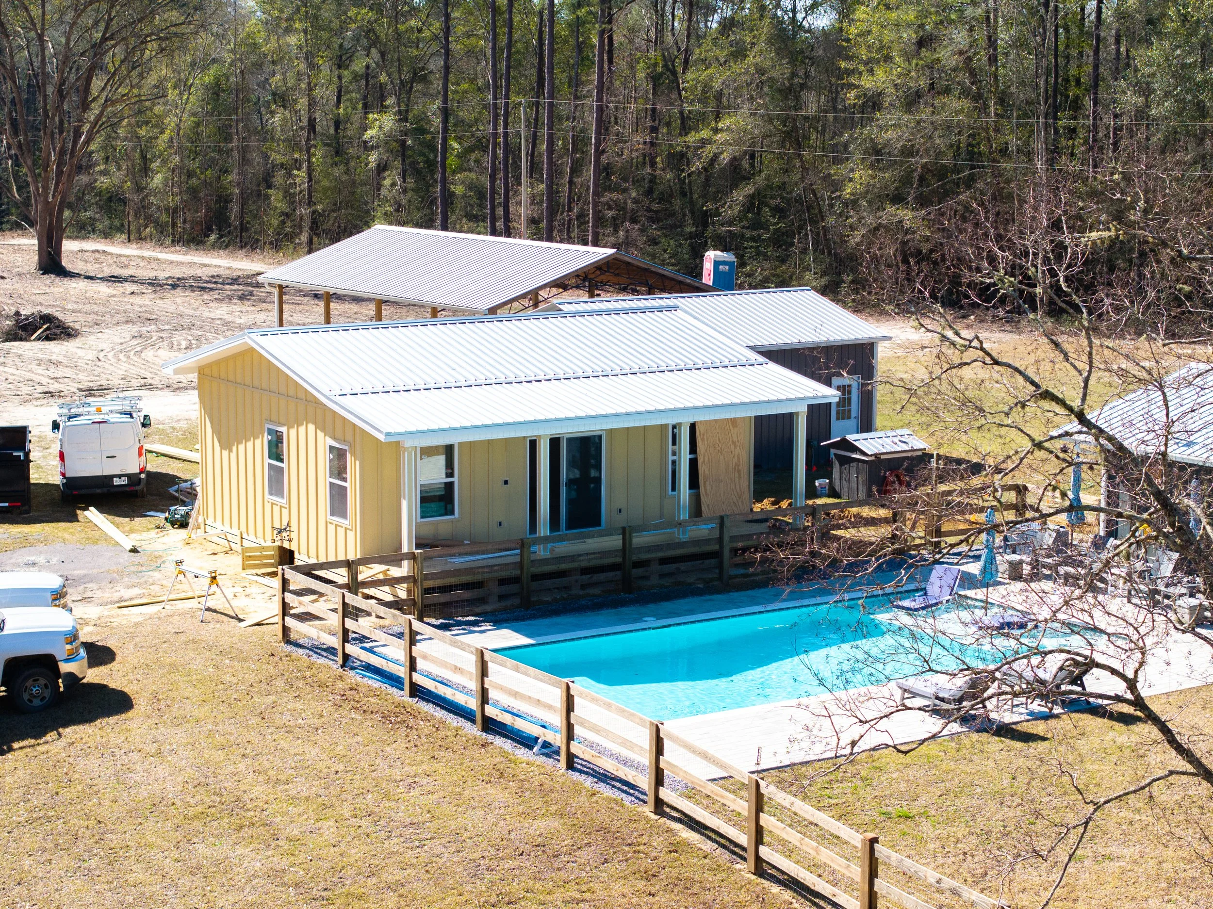 A house with a metal roof next to a swimming pool, surrounded by trees and a yard, with a clear sky above.