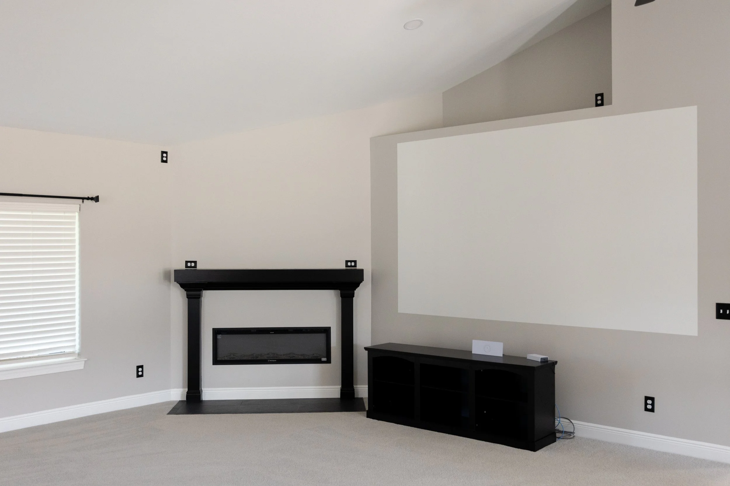 Empty living room with white walls, beige carpet, a window with blinds, and a black fireplace mantle surrounded by multiple electrical outlets.