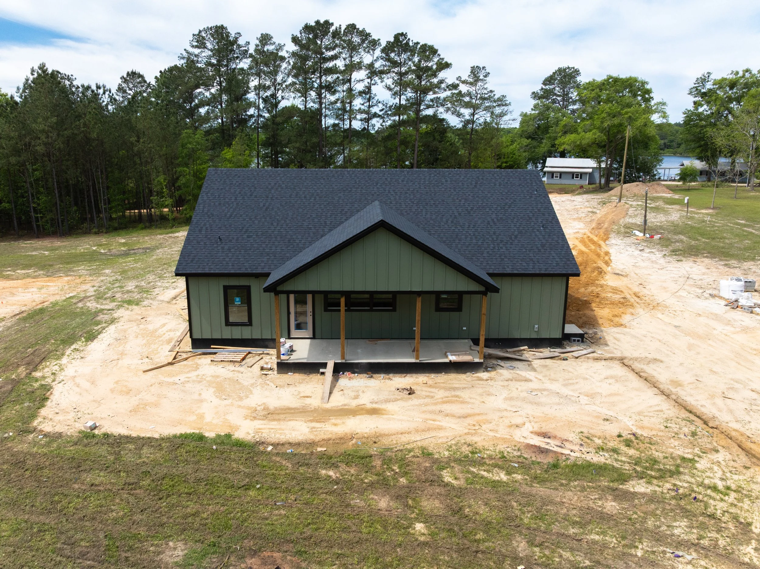 A house under construction with a black shingle roof and green siding, surrounded by dirt and grass, with trees and other houses in the background.