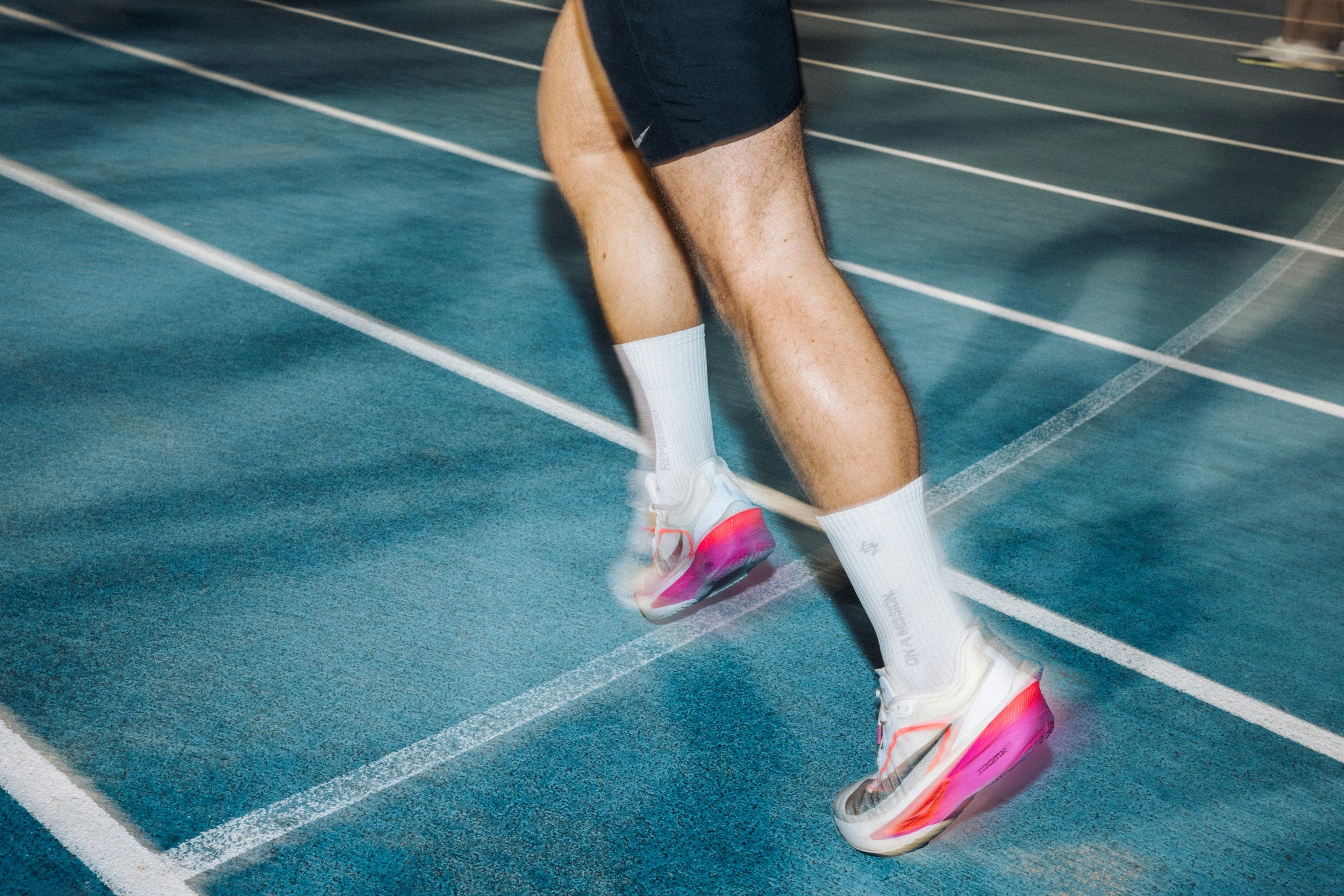 Close-up of a runner's legs on a blue indoor track, wearing white and pink running shoes, white socks, black shorts, and preparing to start a race.