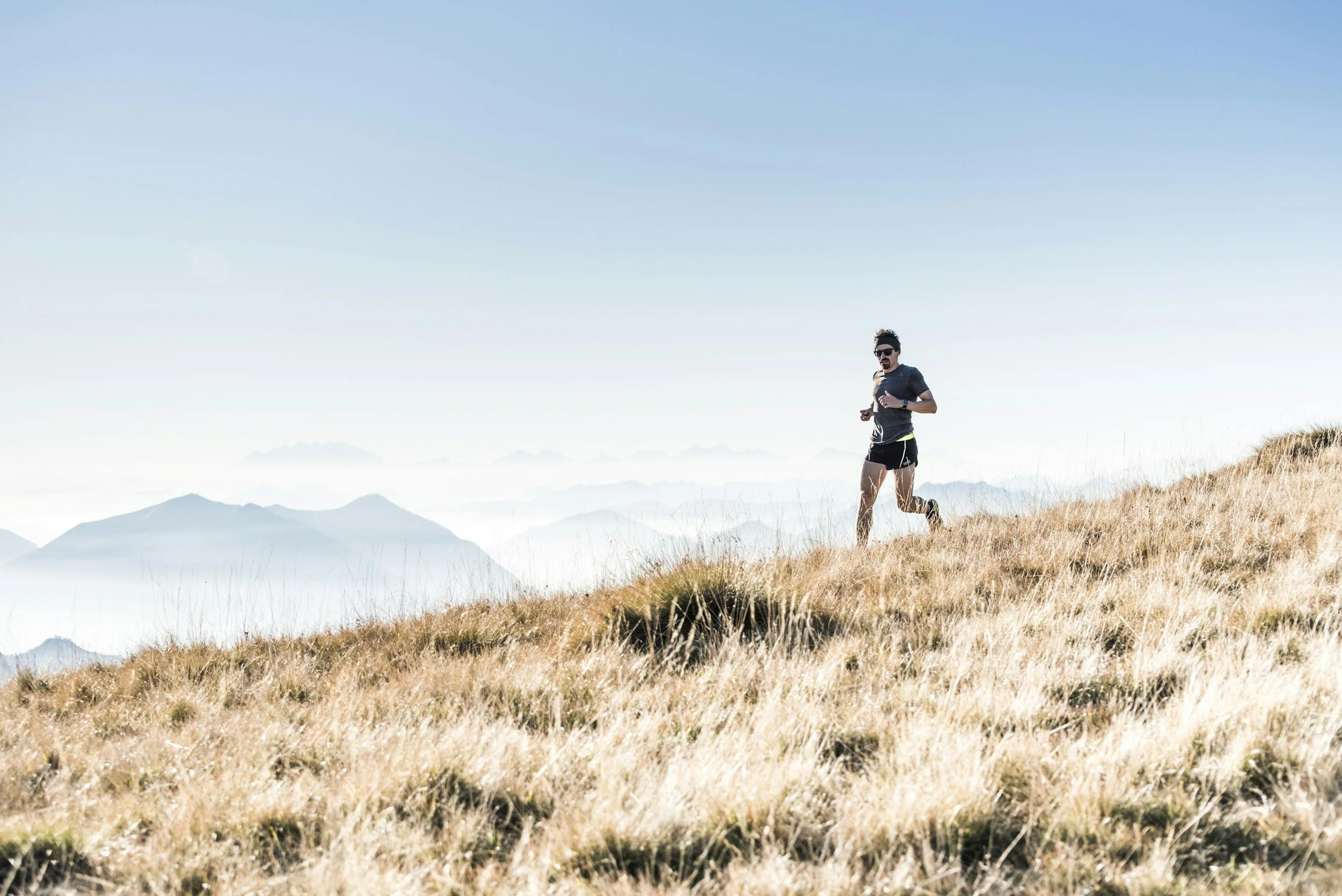 A man running on a grassy hillside with mountains in the background under a clear blue sky.