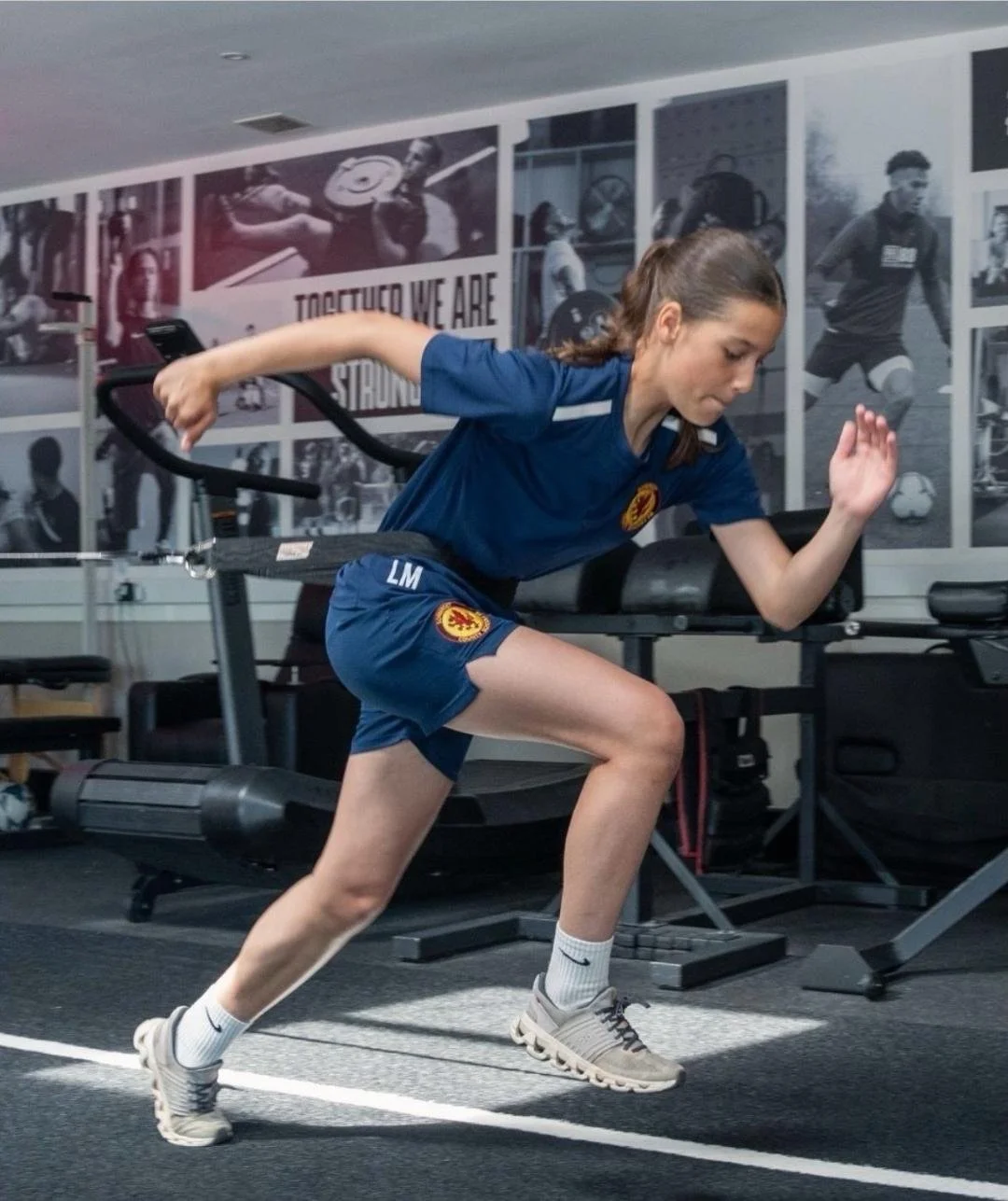 A young woman in sportswear performing a running exercise on a treadmill in a gym, with black-and-white athletic-themed posters on the wall in the background.