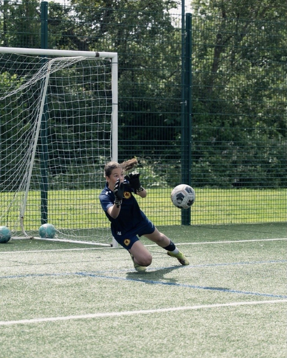 A young girl in a soccer uniform, wearing gloves, is in the process of saving a soccer ball from going into the goal on a soccer field.