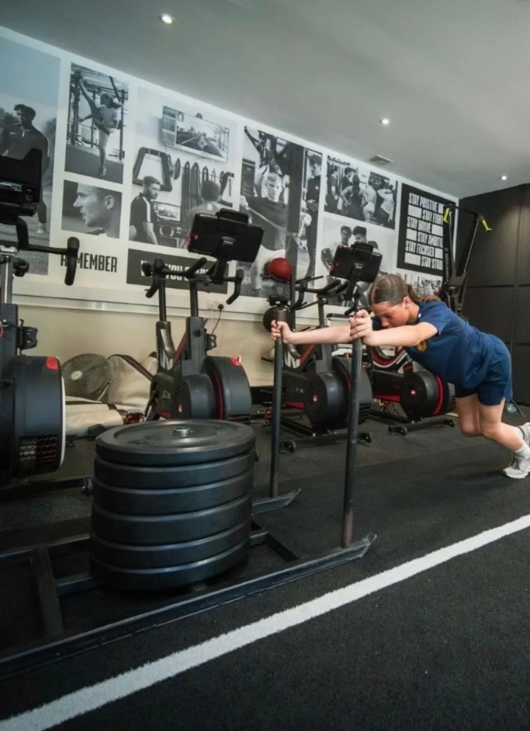 A young girl is pushing a sled in a gym with exercise equipment and black-and-white photos on the wall.