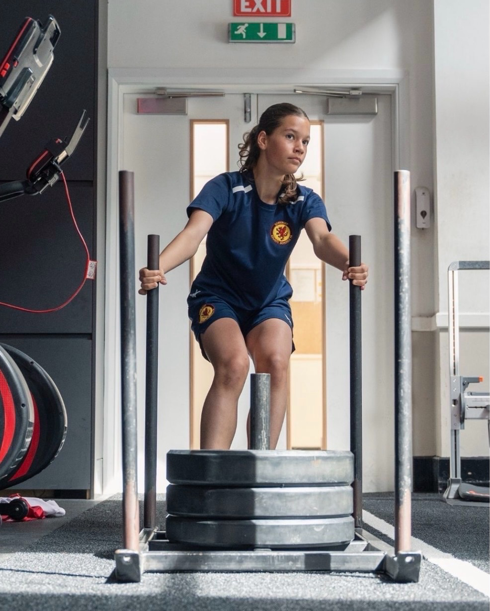 Young woman in a navy sports uniform pushing a weighted sled at a gym.