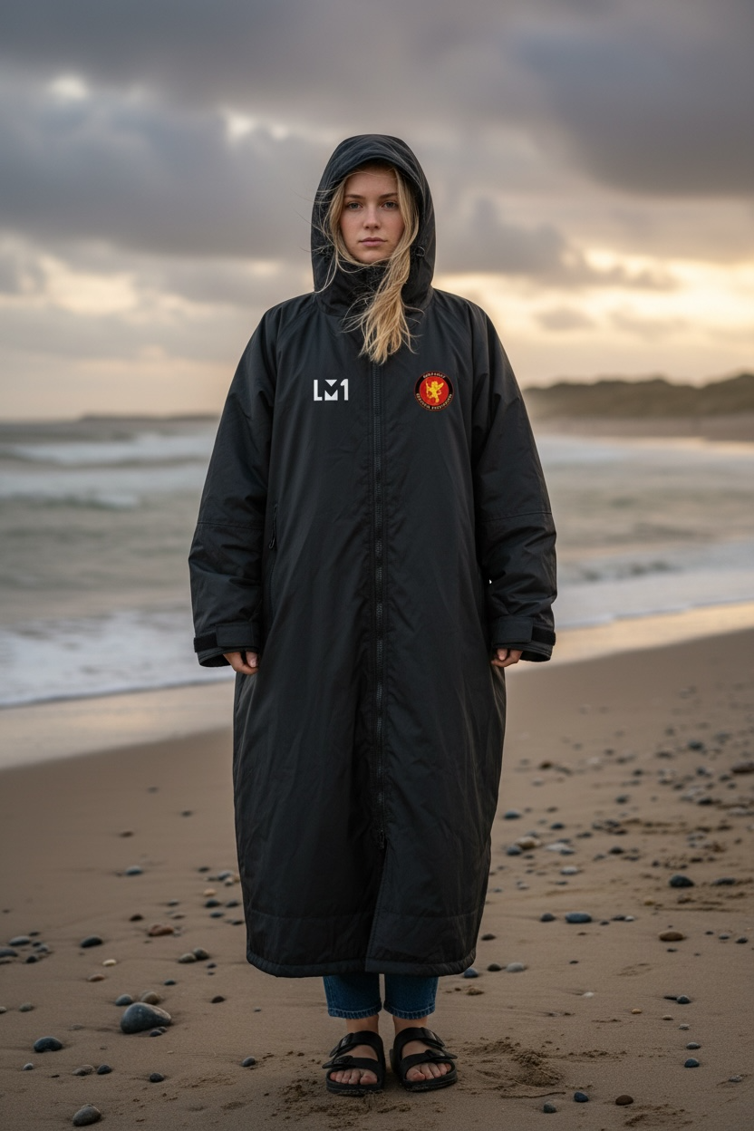 Young woman standing on a beach wearing a long black hooded raincoat, with a cloudy sky and ocean in the background.