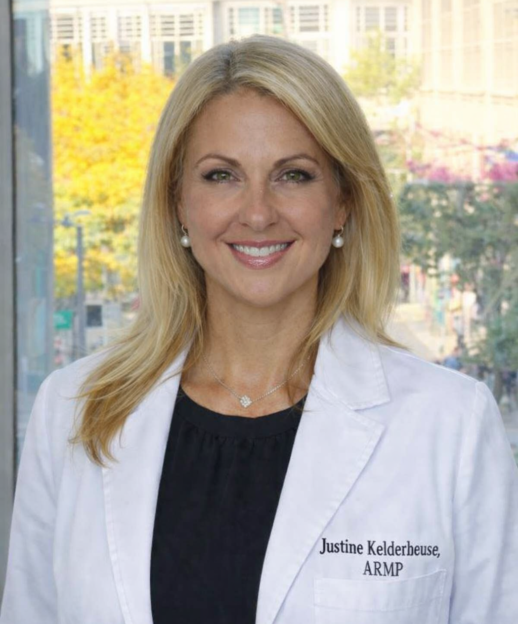 A woman with blonde hair wearing a white medical coat, black top, pearl earrings, and a necklace, standing in front of a window with a city view.