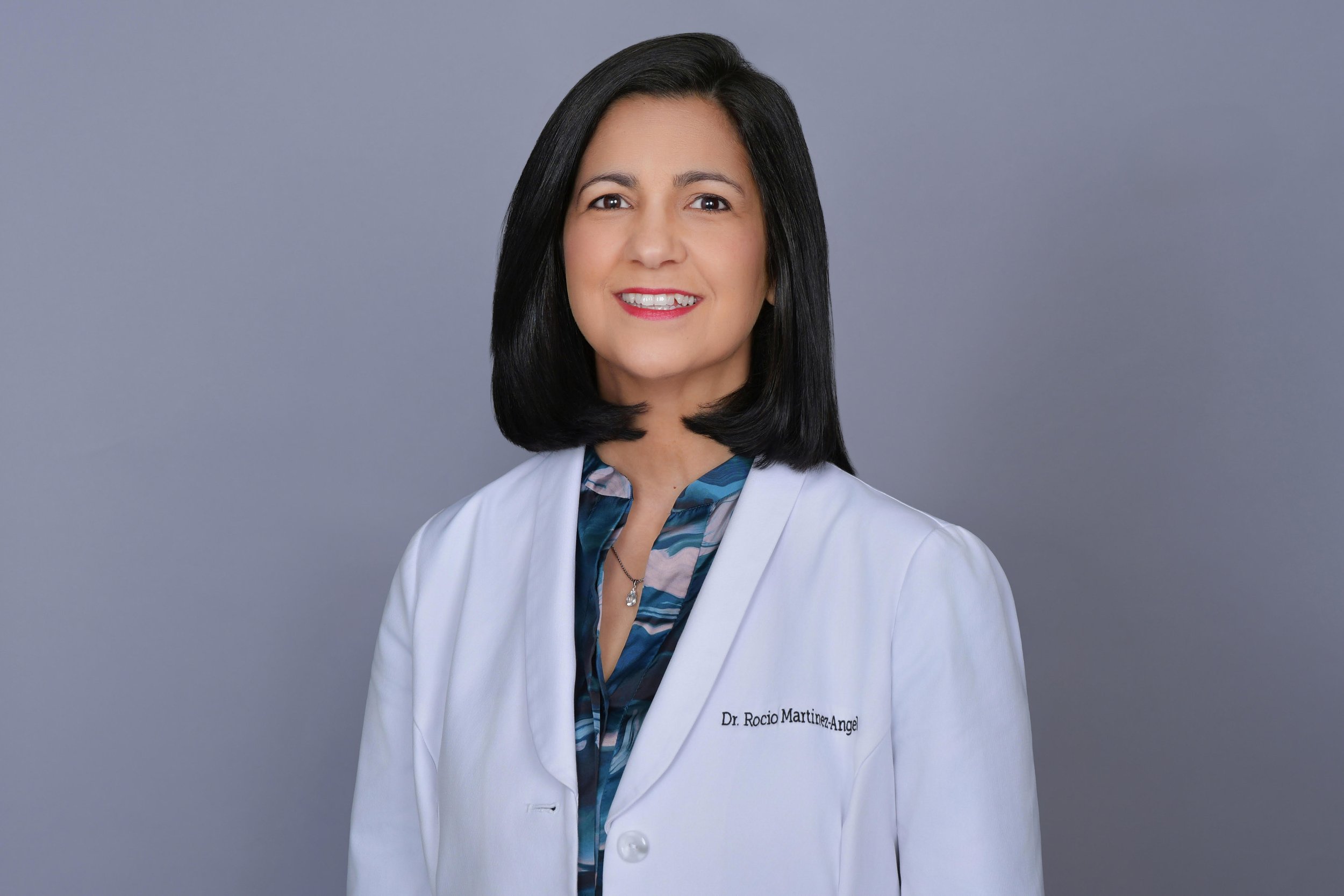 A woman with shoulder-length black hair wearing a white lab coat with the name tag 'Dr. Rocio Martinez-Angel' and a blue patterned blouse underneath, standing against a plain gray background.