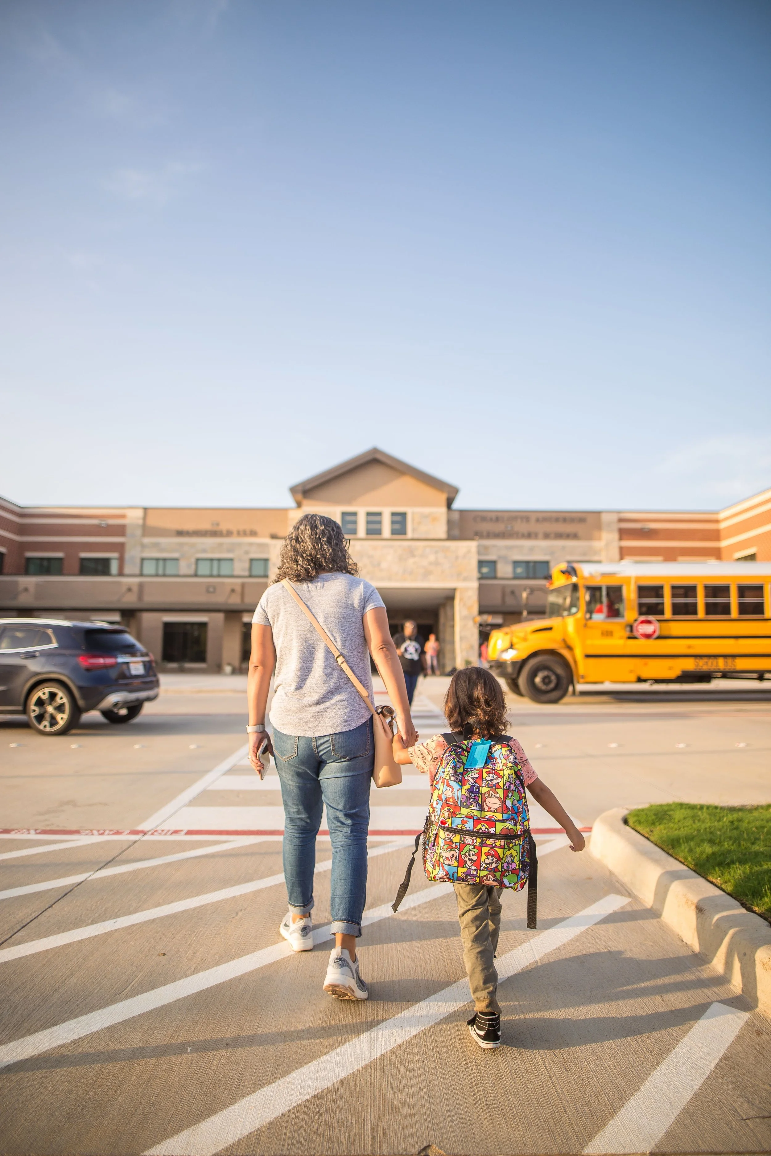 A woman walking with a young girl, holding hands in front of an elementary school, with cars and a yellow school bus nearby, in the late afternoon sunlight.