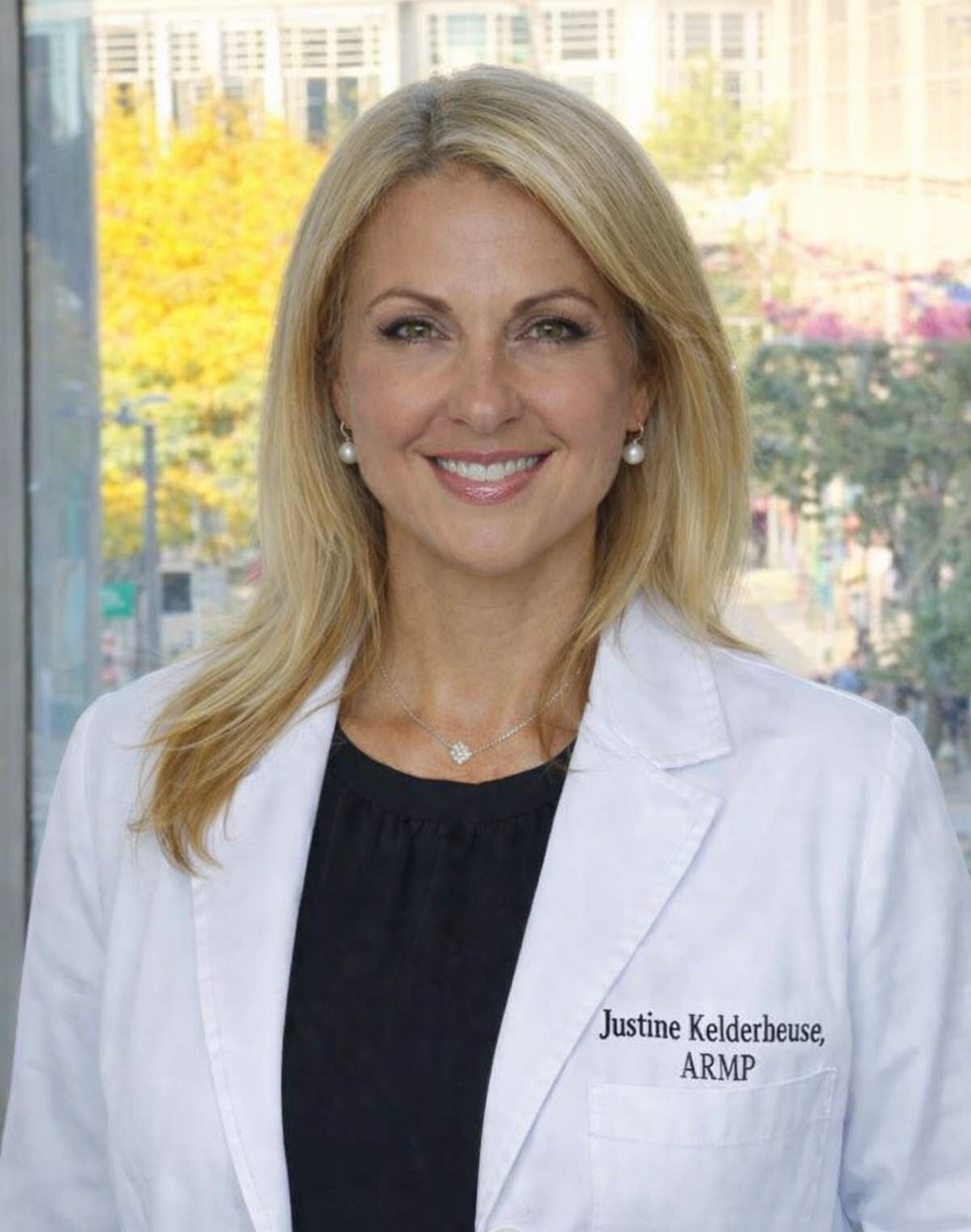A woman with blonde hair wearing a white medical coat and a black top, standing indoors near a window with a cityscape background, smiling at the camera.