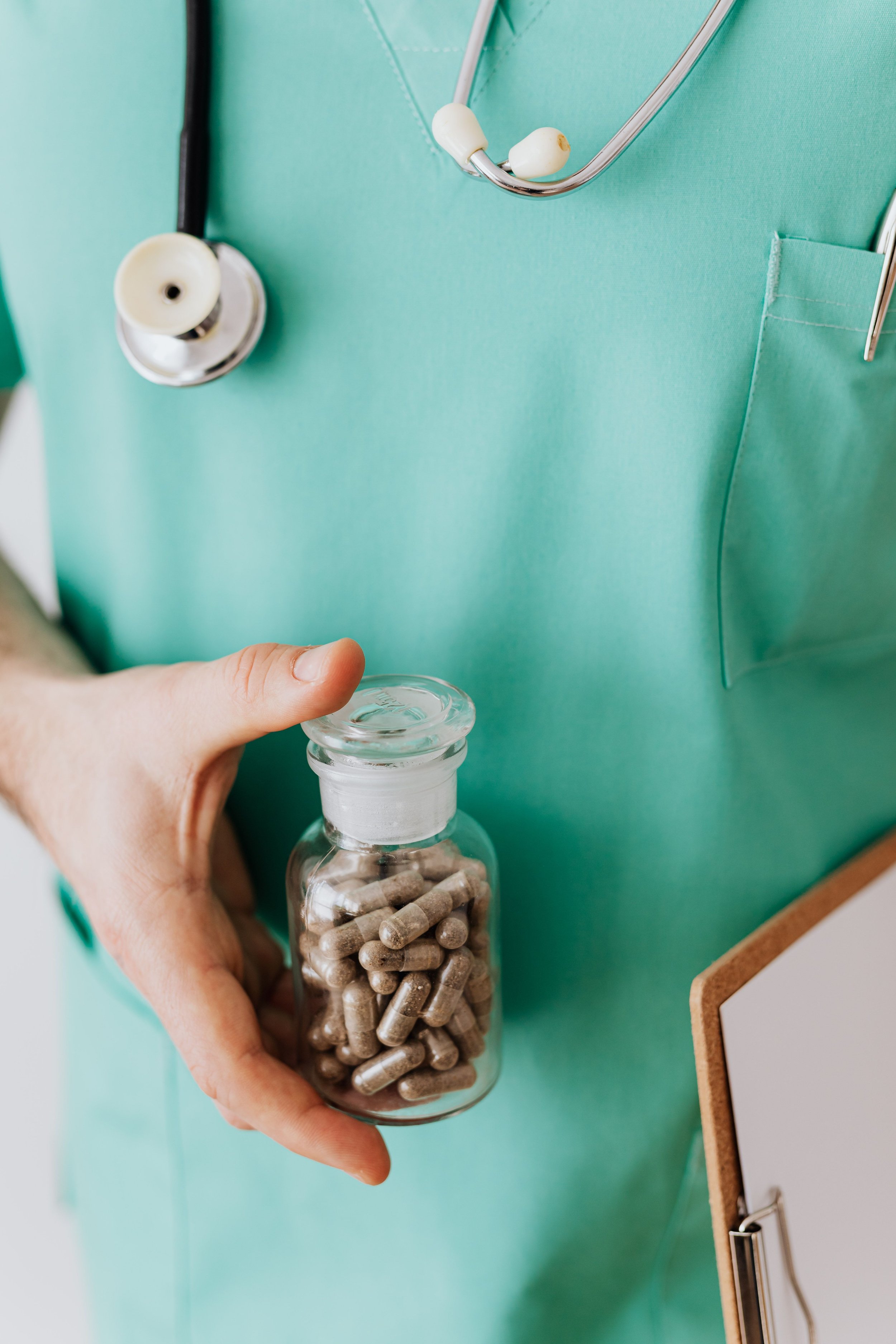 A person in a green medical uniform holding a small bottle of pills and a stethoscope hanging on the chest.