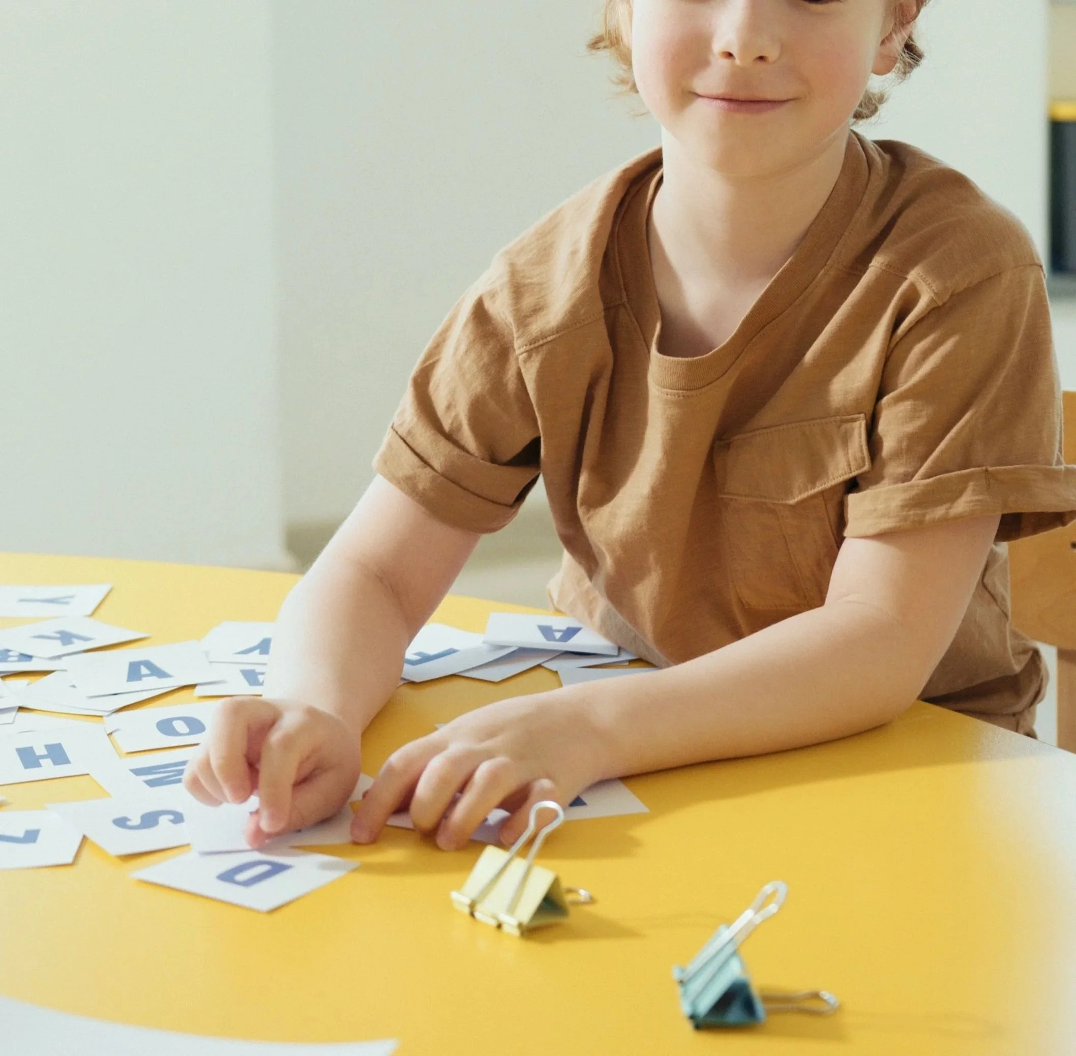 A young boy with red hair and a tan shirt is sitting at a yellow table, looking down and working with alphabet flashcards and paper clips.