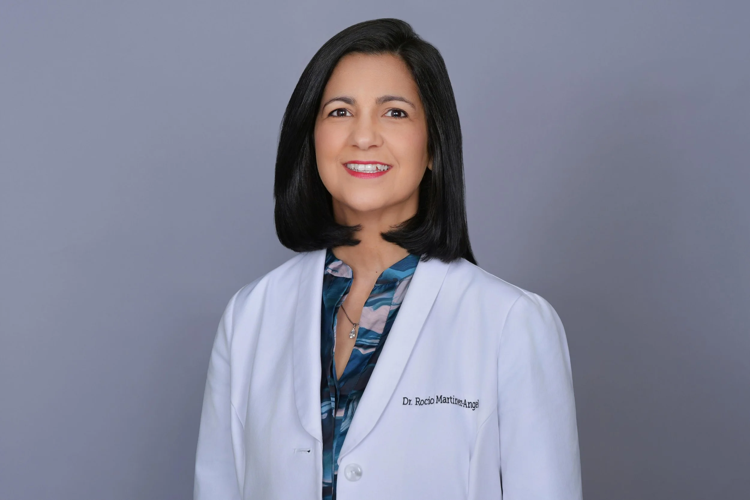 Portrait of a woman with shoulder-length black hair wearing a white lab coat with a name tag that reads 'Dr. Rocio Martinez-Angel,' a blue patterned blouse, and a necklace, standing against a neutral gray background.