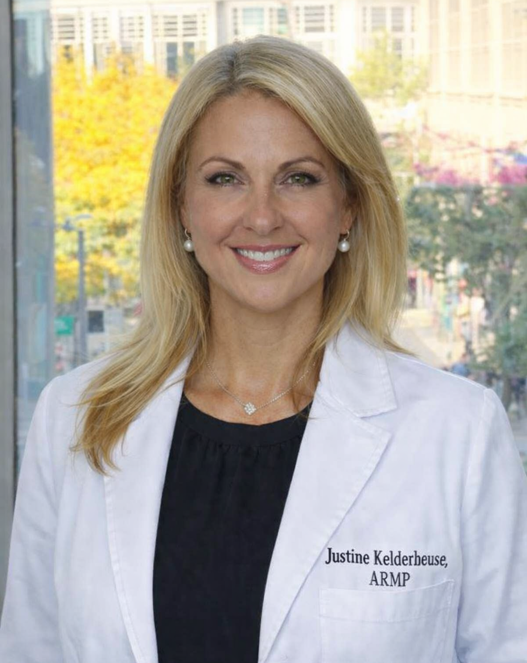A woman with blonde hair wearing a white coat with "Justine Kelderheuse, ARMP" embroidered on it. She is smiling and wearing pearl earrings and a necklace, standing indoors with a cityscape and trees visible through a window behind her.