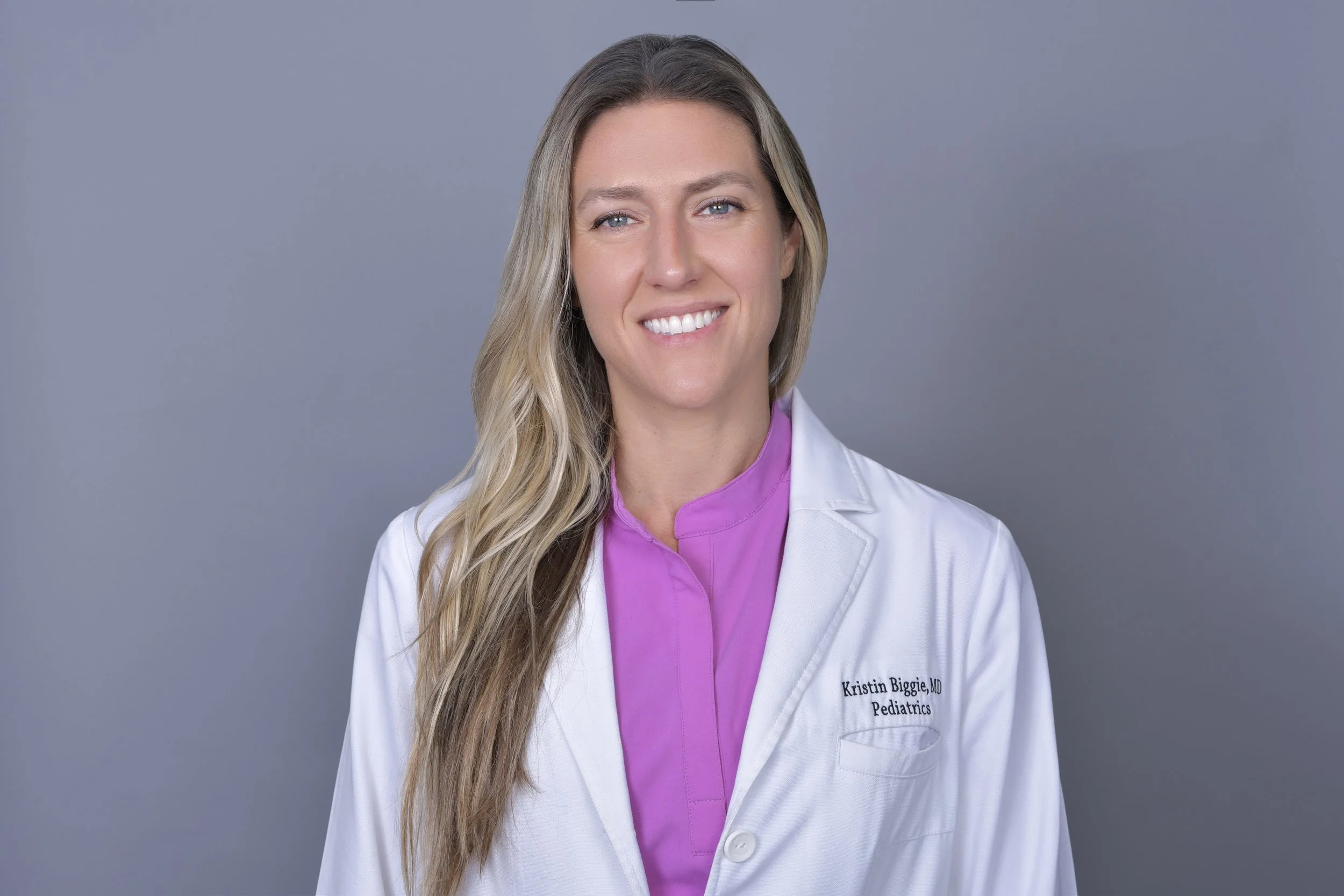 Portrait of a woman with long blond hair, wearing a white coat over a purple shirt, smiling against a gray background.