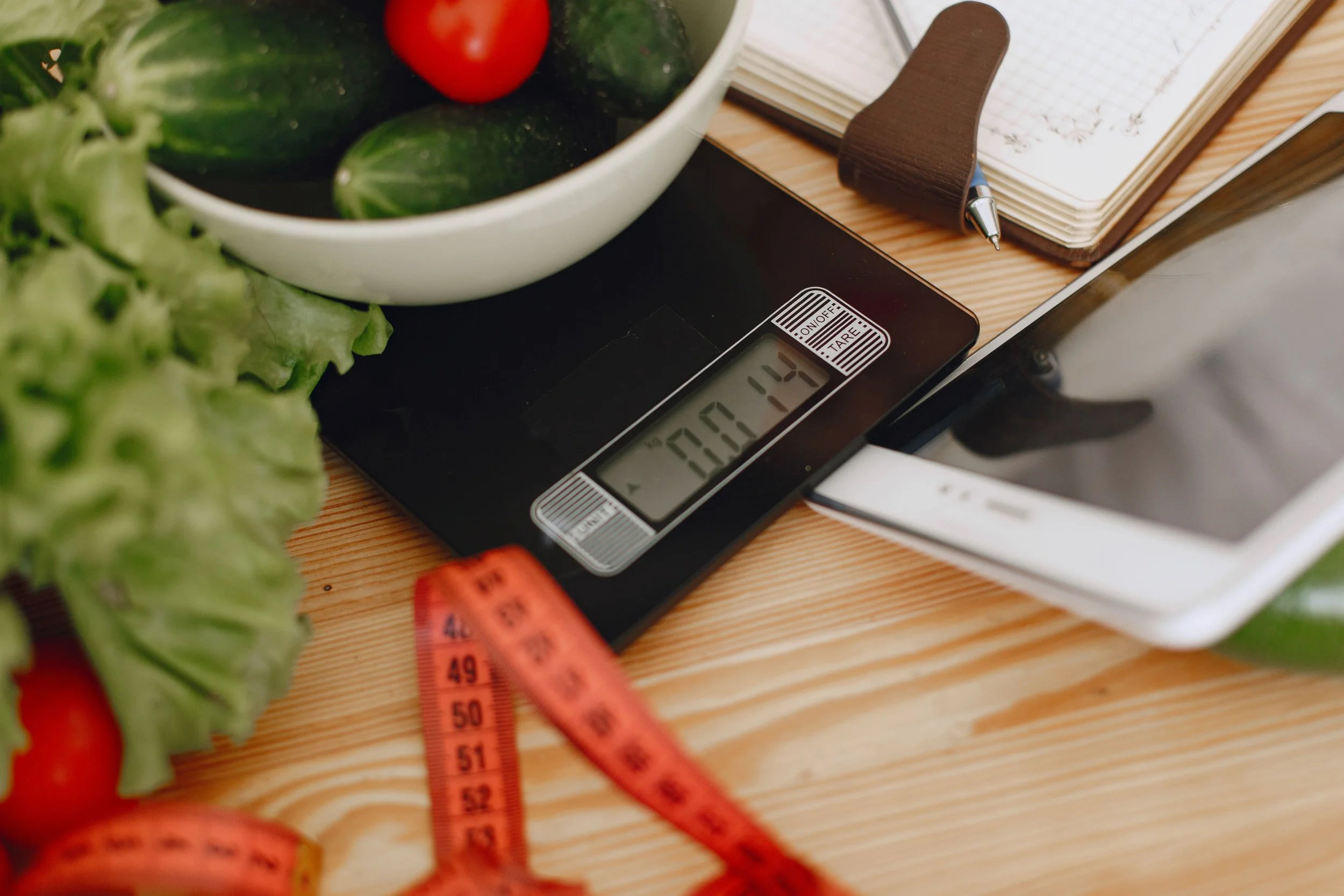 A kitchen scale on a wooden countertop with a bowl of vegetables and a pink measuring tape nearby.