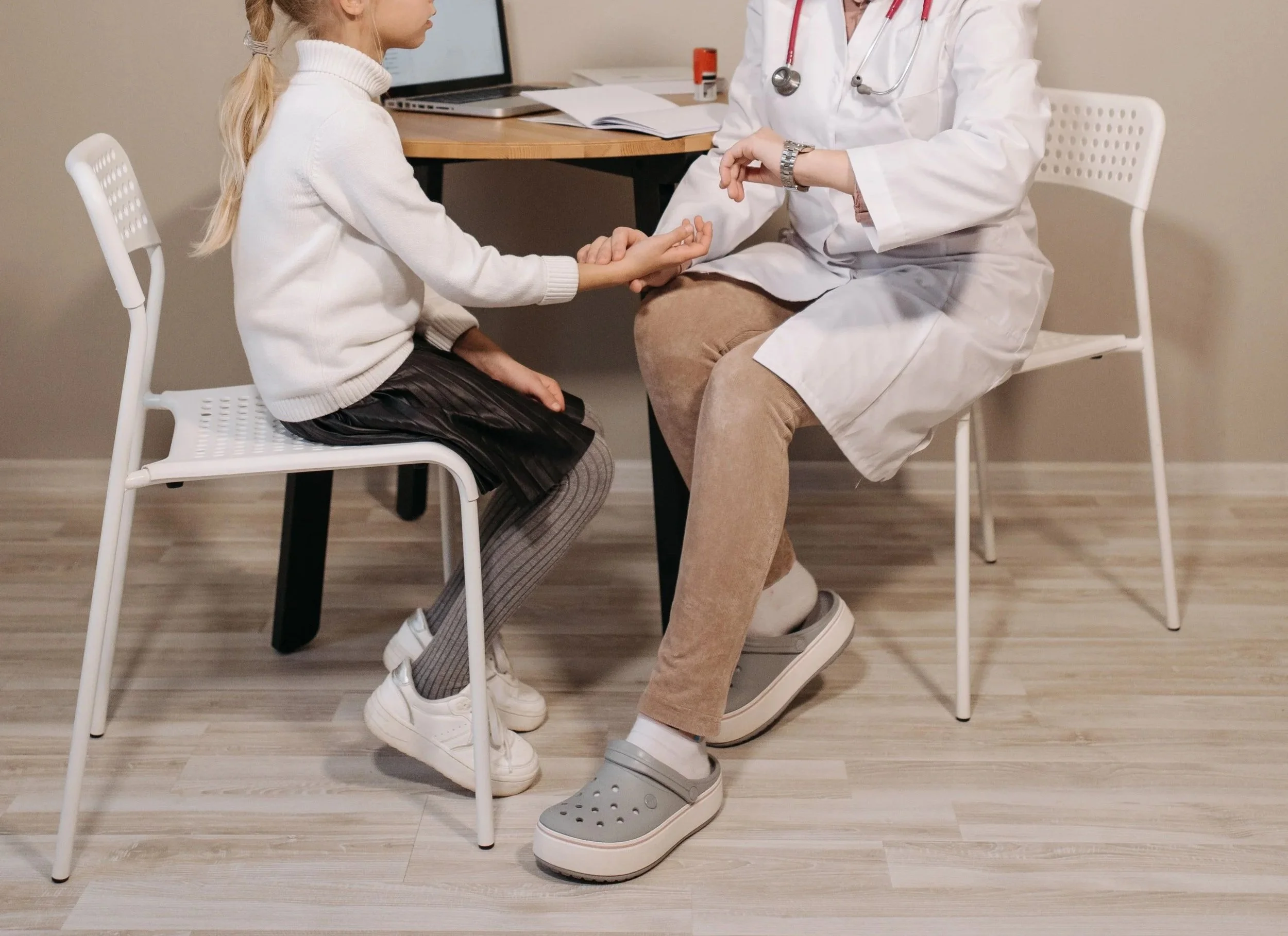 A young girl with blonde hair sitting in a white chair at a doctor's office, holding hands with a female doctor who is wearing a white coat and grey sneakers. The doctor is looking at her wristwatch. There is a laptop, some papers, and a prescription bottle on a small table behind them. The wall behind them is beige with a black clock showing 3:26.