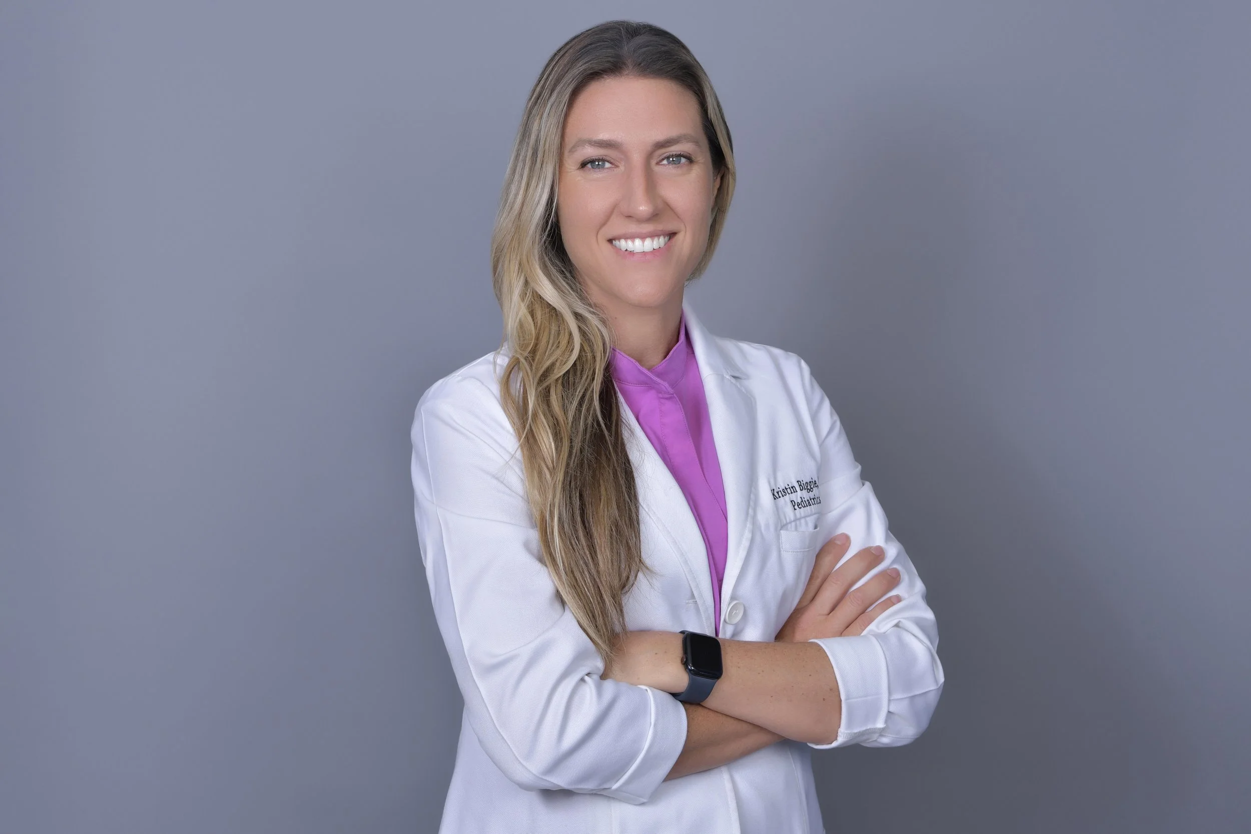 Portrait of a smiling female pediatrician in a white coat with long wavy blonde hair, standing with arms crossed against a plain gray background.