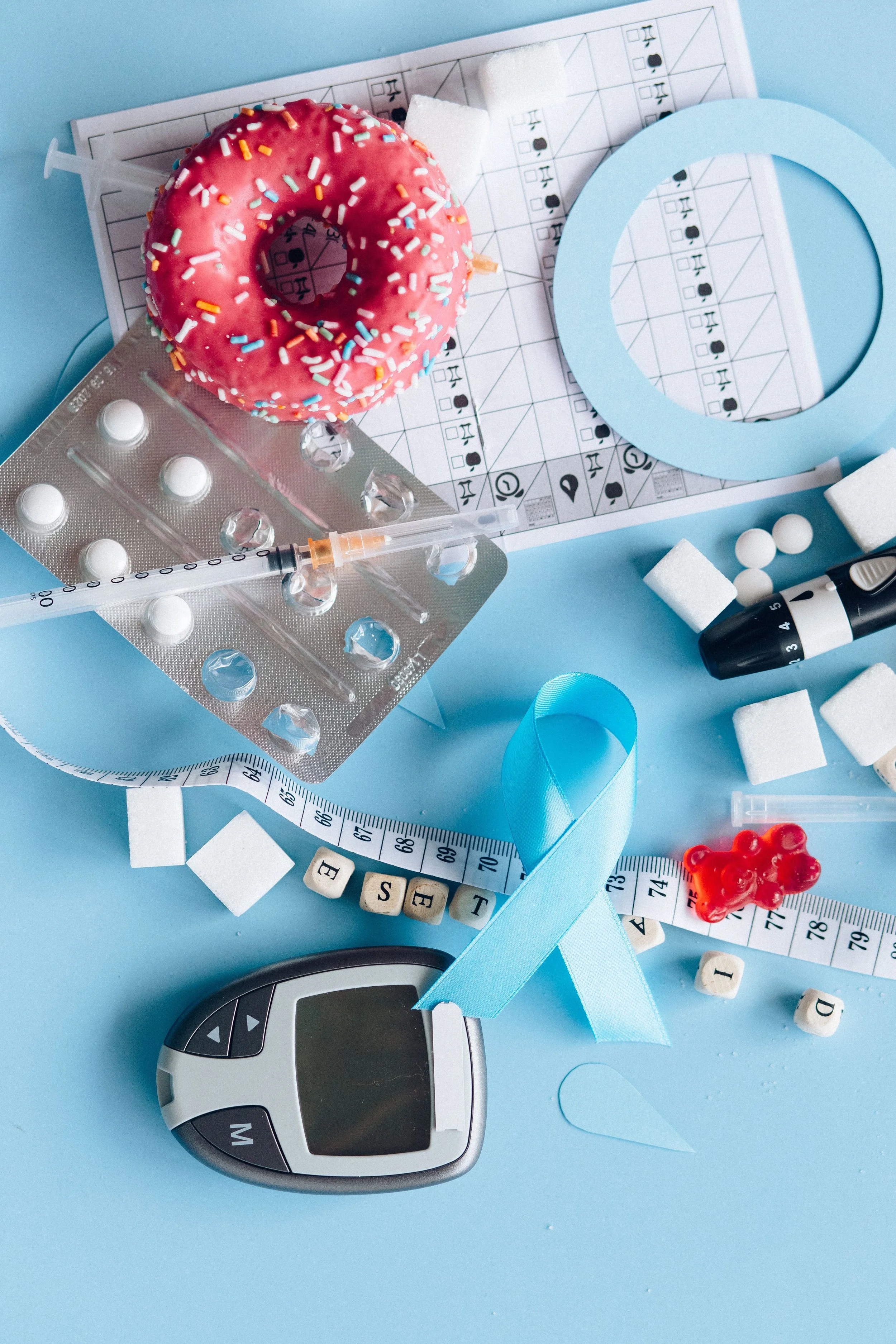 Medical scene featuring a pink frosted donut with sprinkles, a syringe, white pills, medication blister pack, a blue circular object, a black and white timer, white sugar cubes, letter and number blocks, blue ribbon, and a tape measure on a light blue background.