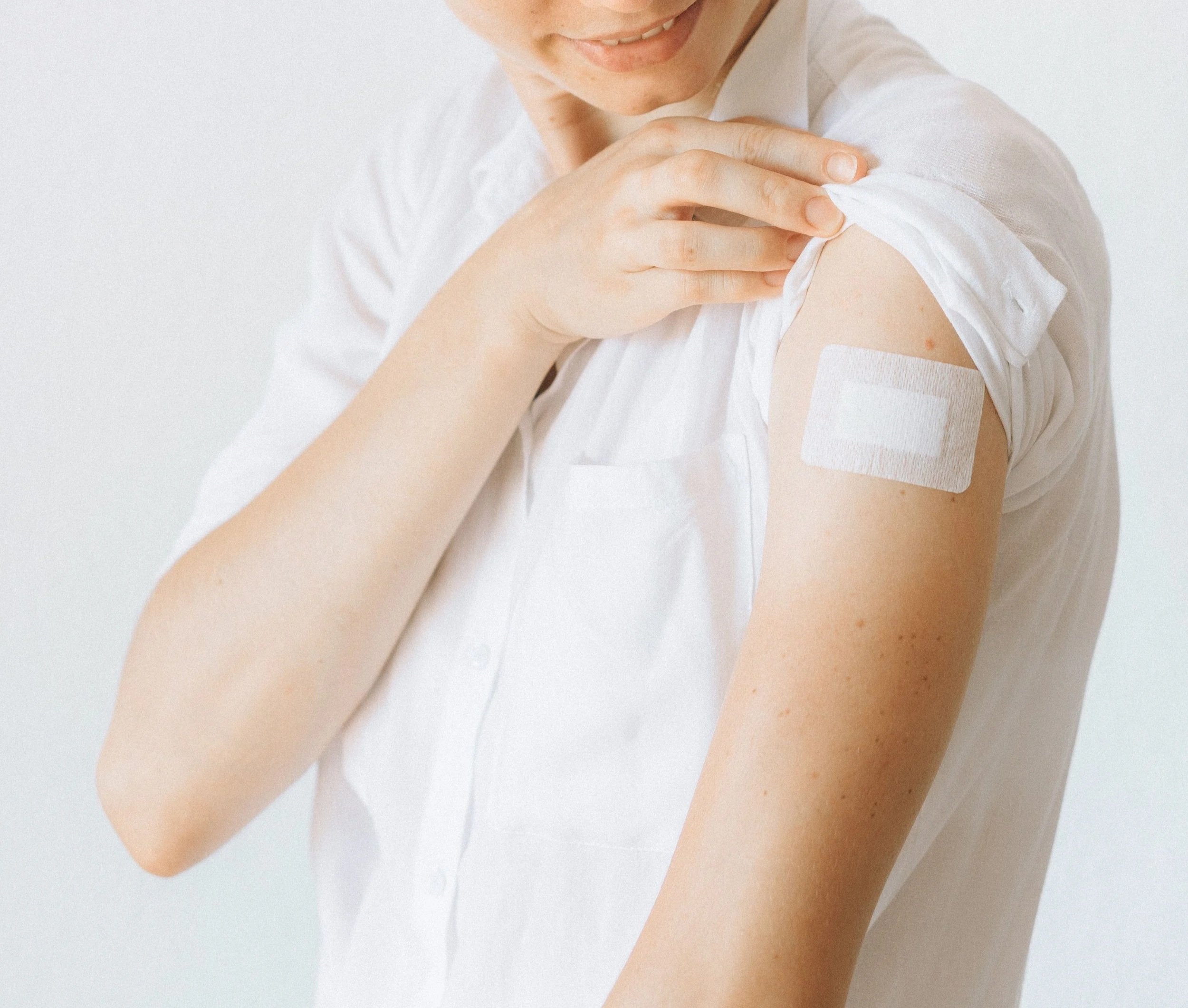 A woman with blonde hair is looking down and smiling, her eyes closed. She is wearing a white shirt and has a bandage on her upper arm, where she just received a vaccination or shot.