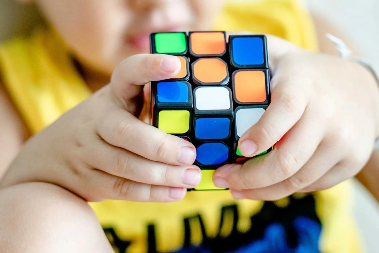 A person holding a Rubik's cube with one hand, with part of their face visible in the background and their arm resting on their knee, wearing a yellow shirt.