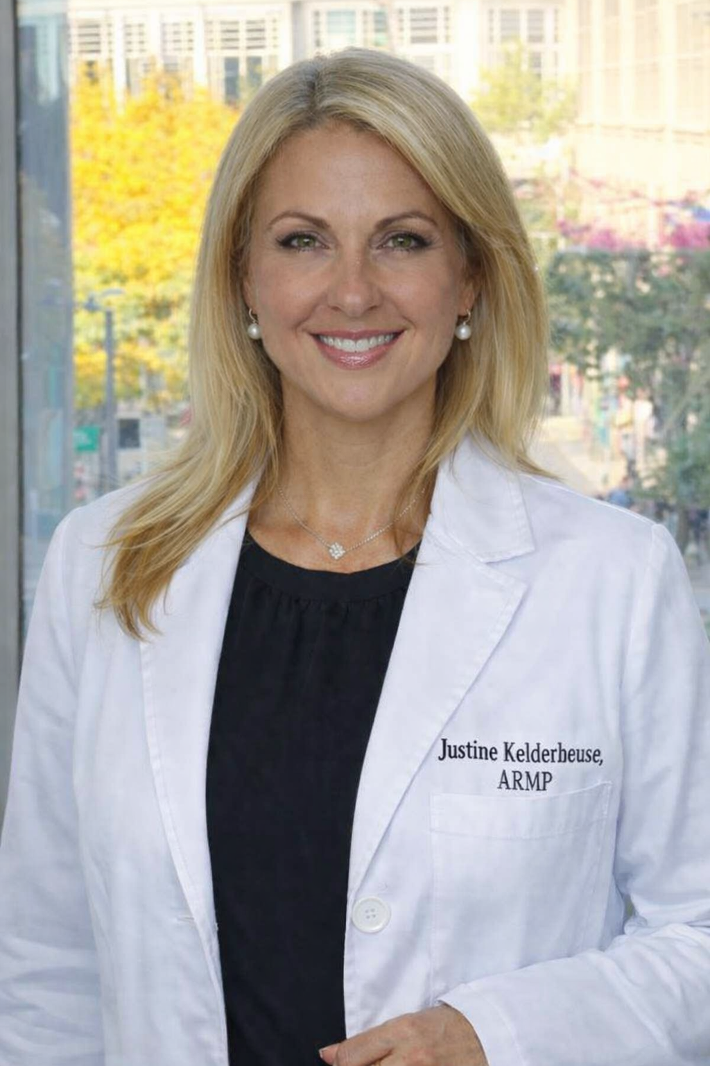 A woman with blonde hair wearing a white medical coat with the name 'Justine Kelderhouse, ARMP' embroidered on it, standing indoors with a cityscape visible through a window behind her.