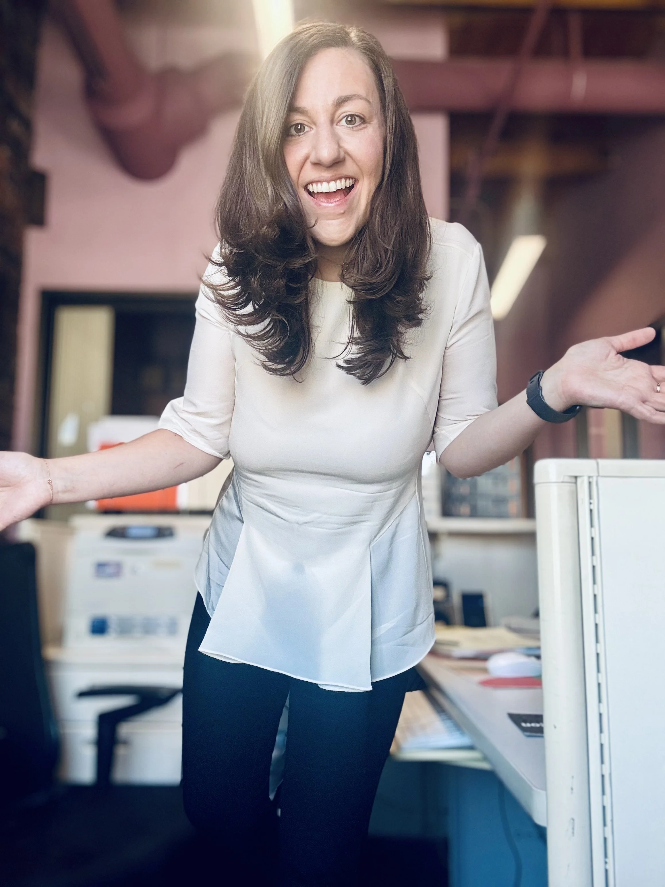 A woman with long brown hair smiling and gesturing with her hands in an office environment with printers and desks.