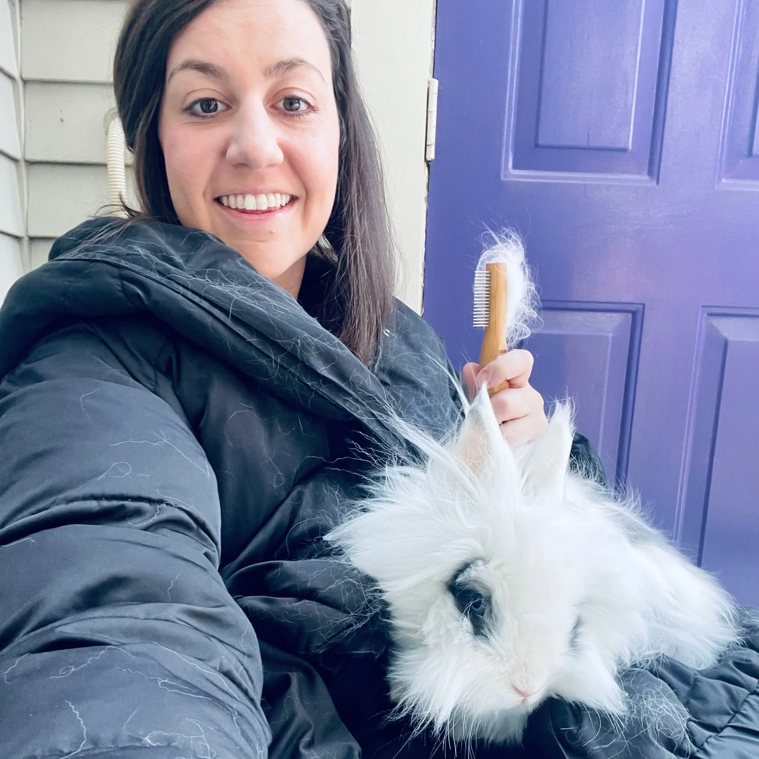 A smiling woman with long dark hair, wearing a black jacket, holding a white fluffy rabbit, standing in front of a purple door.