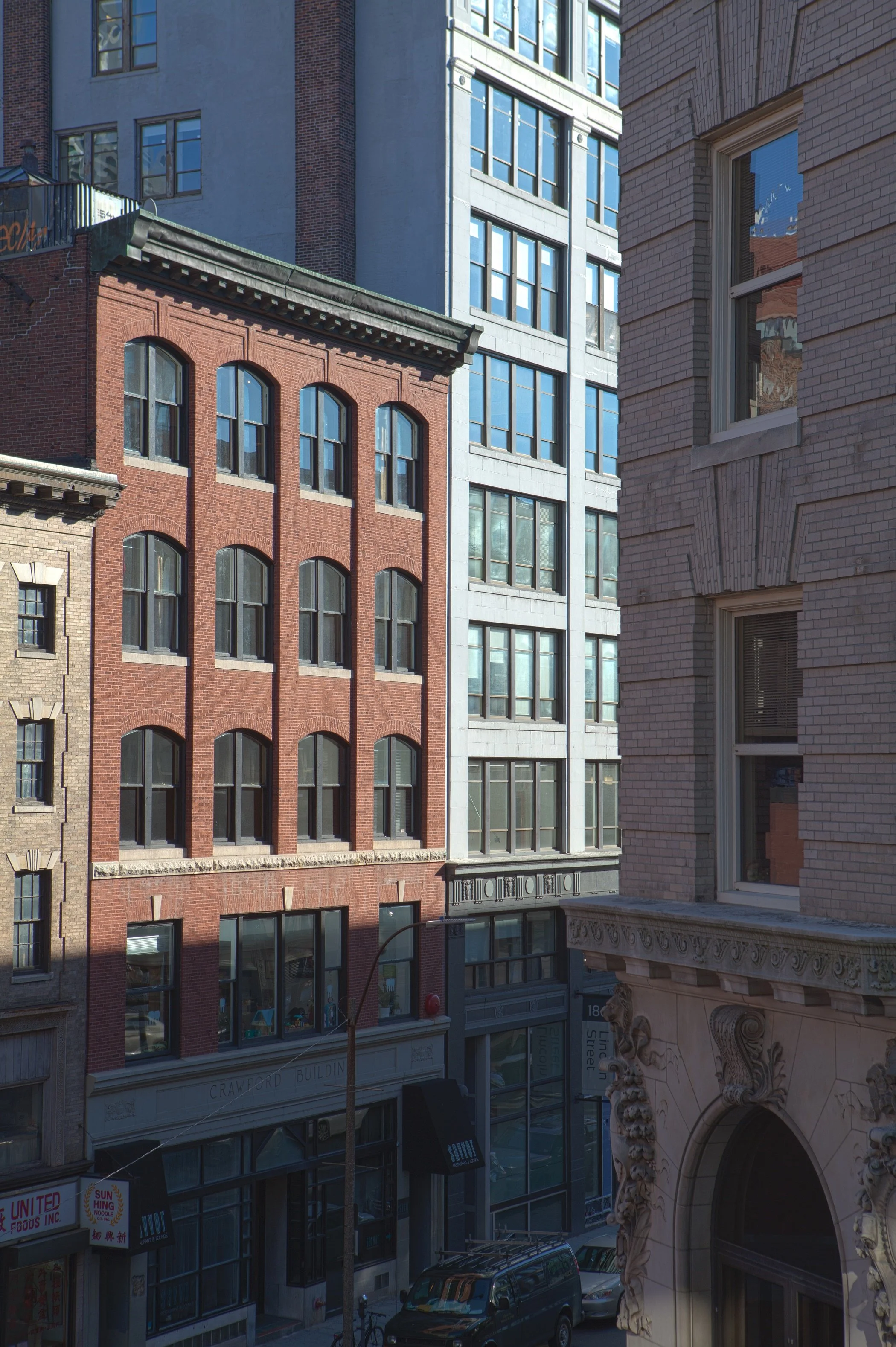 View of multiple multi-story buildings with brick and glass facades on a city street, with parked cars and storefronts at the ground level.
