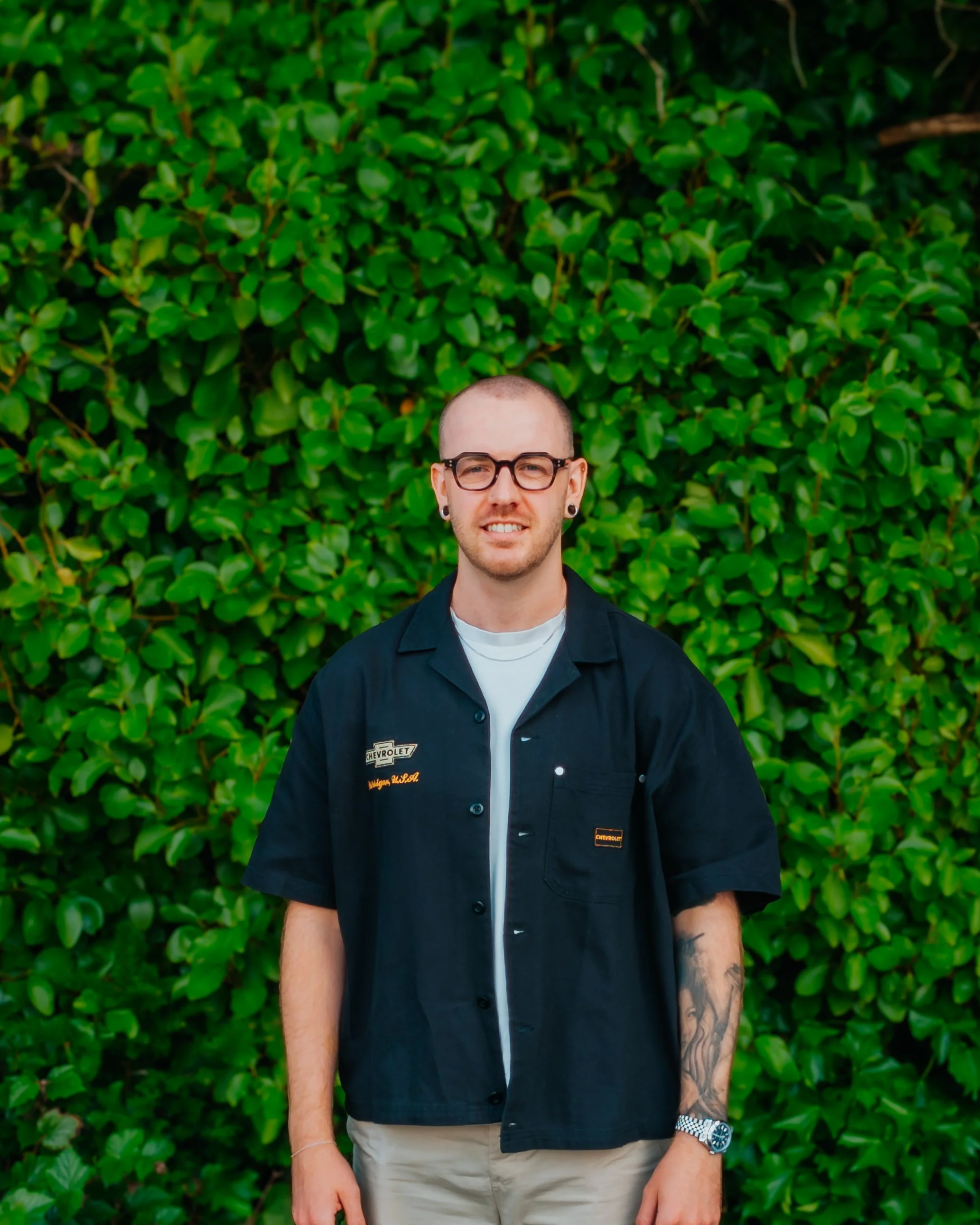 A picture of Jake, the financial adviser with a shaved head and glasses, standing in front of a dense green hedge, wearing a black short-sleeved shirt with a white undershirt and a silver watch.