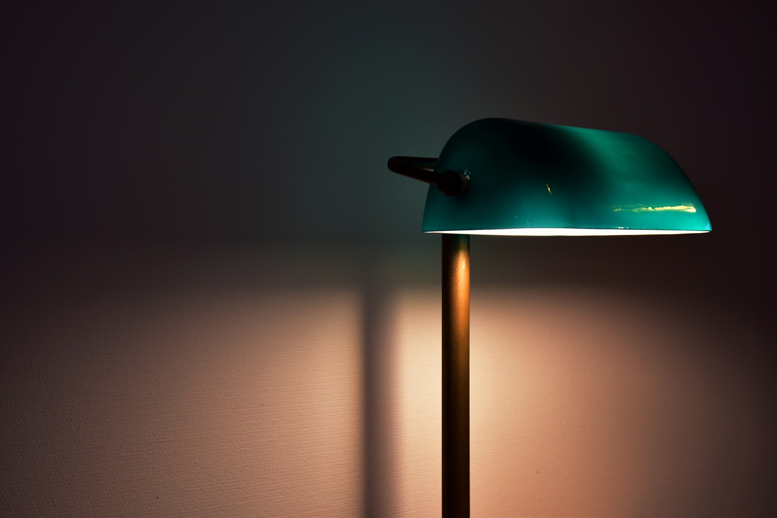 A modern green desk lamp with a wooden stand, turned on and casting a warm light on a wall.