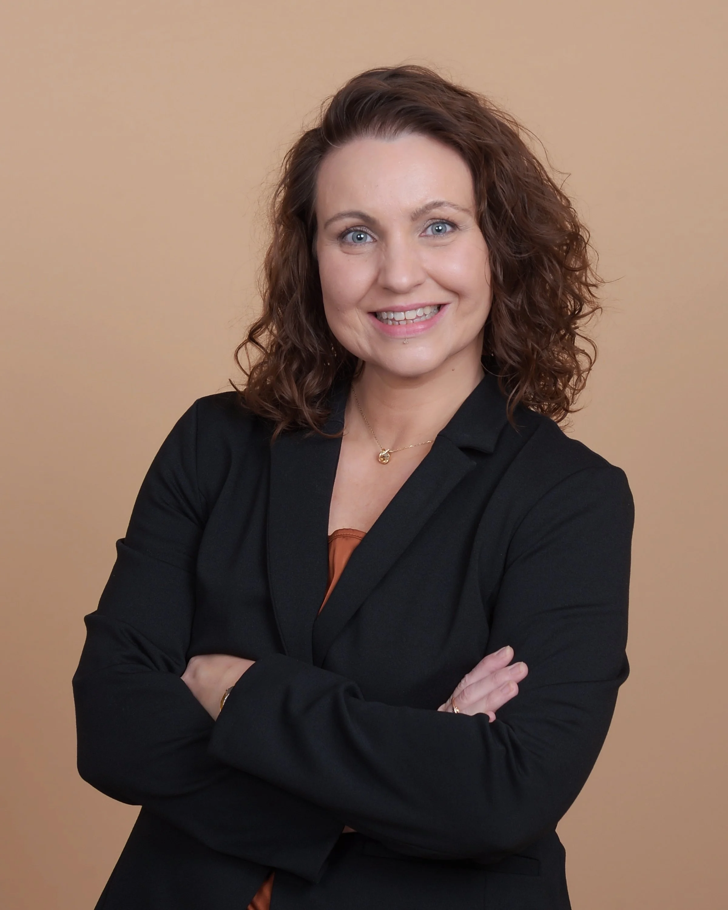 Portrait of a woman with curly brown hair, blue eyes, smiling, wearing a black blazer, crossing her arms, standing against a beige background.