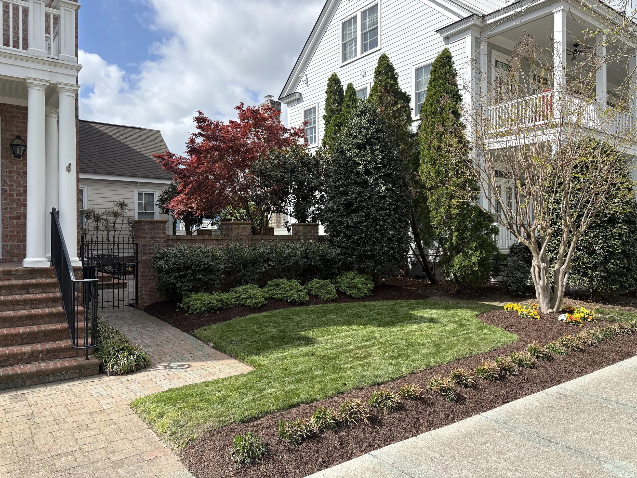 A landscaped backyard with a brick patio, green lawn, well-maintained flower beds, trees including a red-leaved Japanese maple, and a white house with a porch and multiple windows.