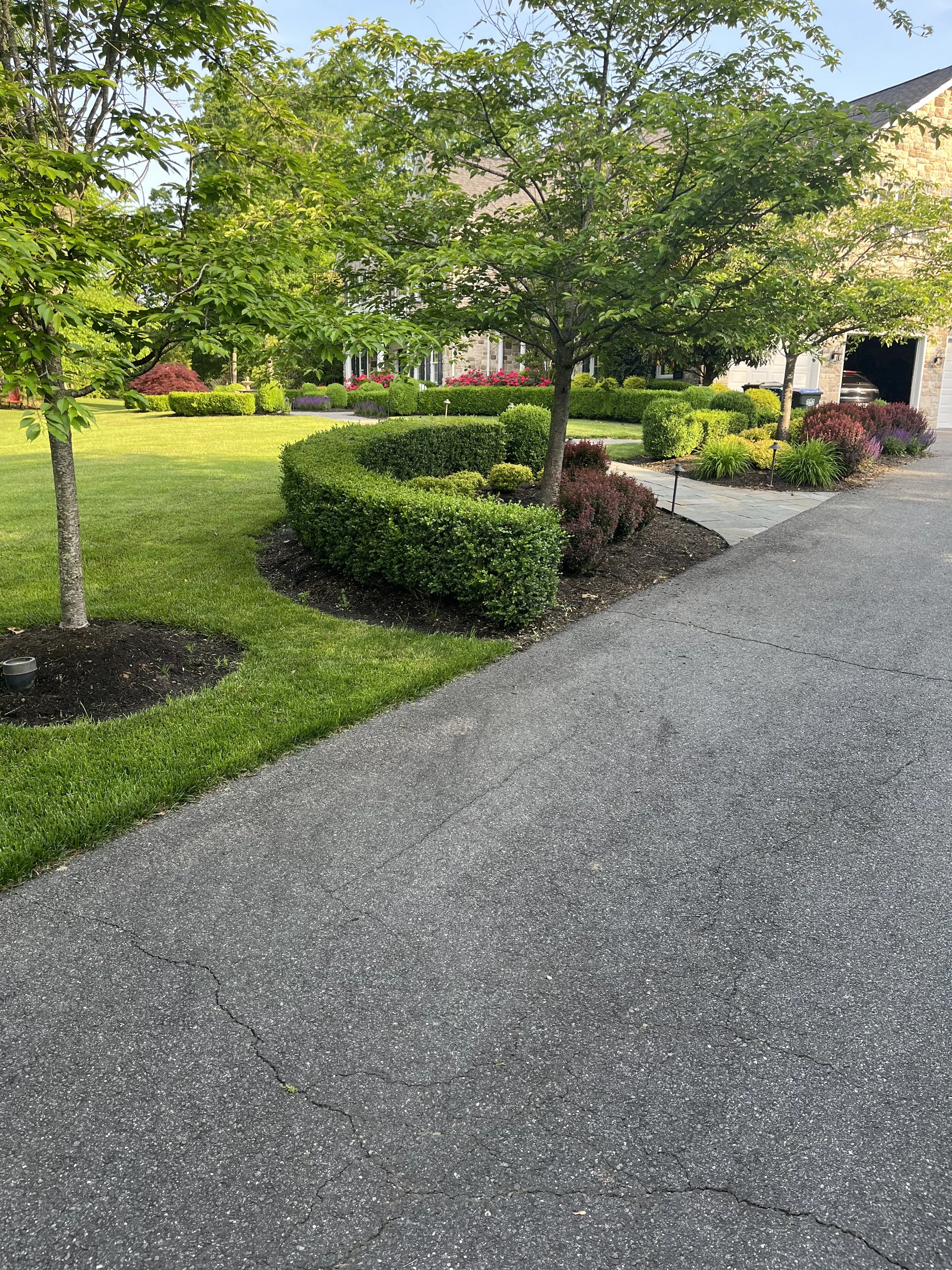 A residential front yard with a green lawn, trees, and well-maintained shrubs. Paved pathway leads to the house, which has a stone exterior and a garage with a car inside. The scene is bright and sunny.