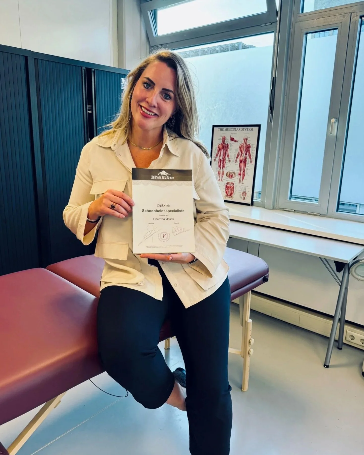 Vrouw glimlachend, zittend op een bankje in een spreekkamer, houdt een diploma vast dat haar kwalificatie als schoonheidsspecialist bevestigt.