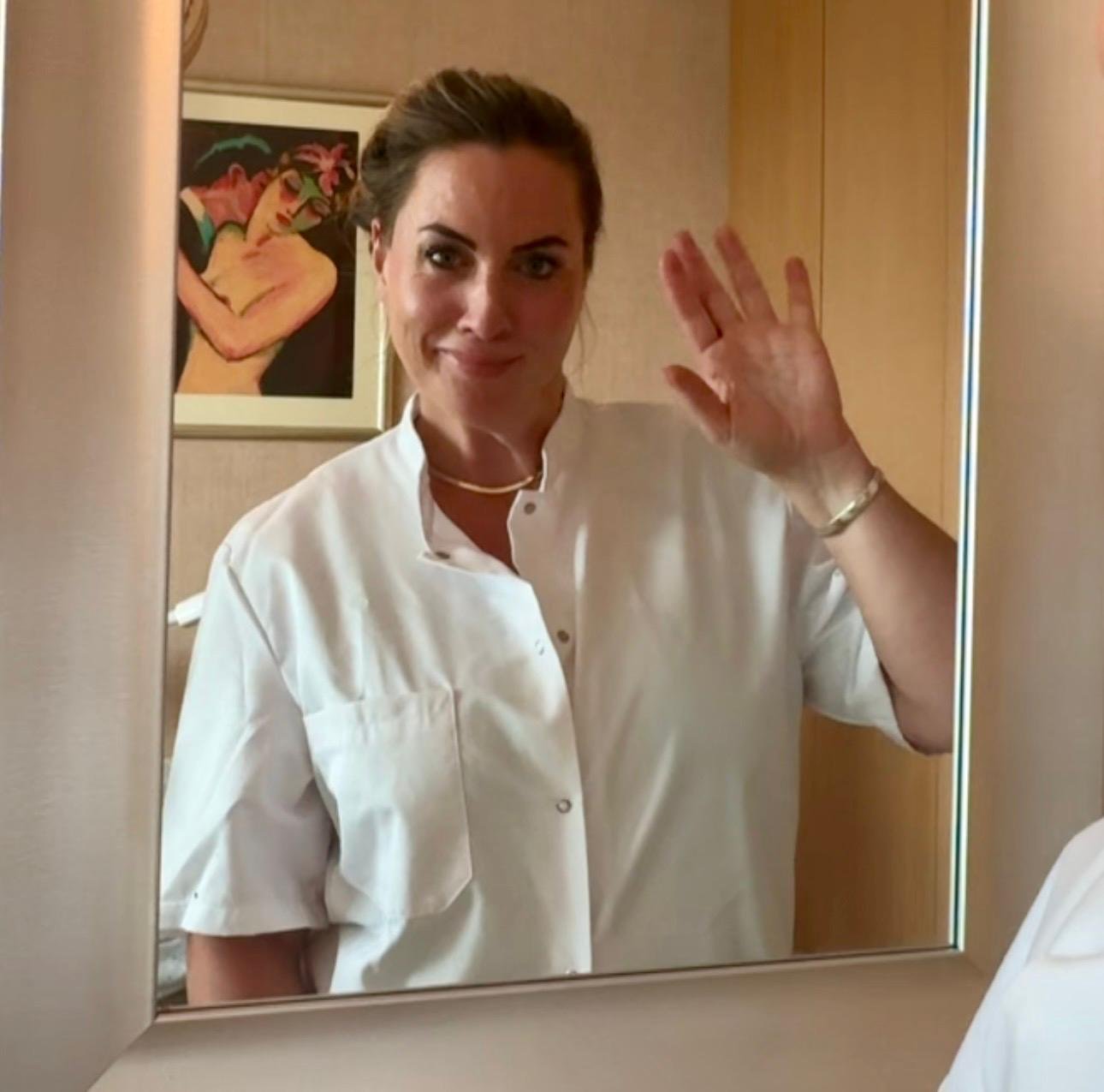 Vrouw in witte ziekenhuisoutfit die naar de camera glimlacht en zwaait, zichtbaar in een spiegel.