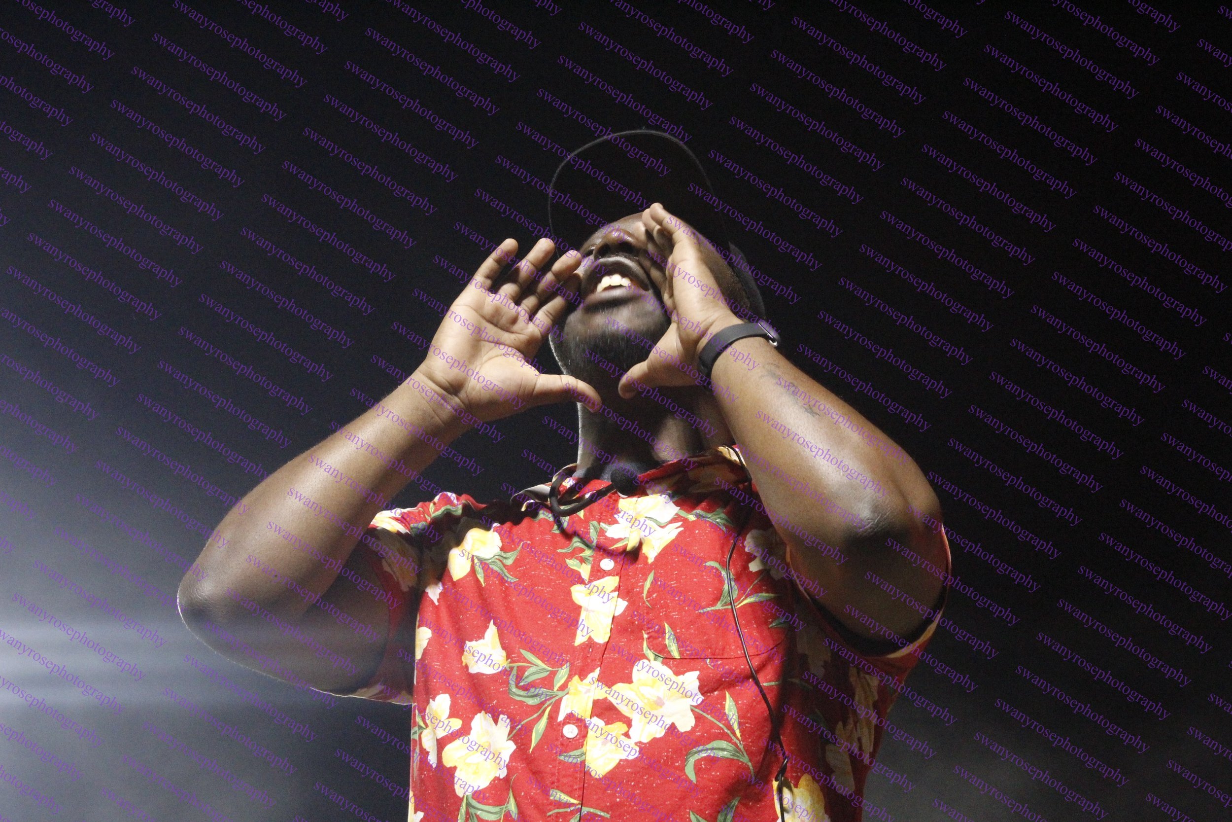 A man laughing with his hands near his face, wearing a red floral shirt, against a dark background.