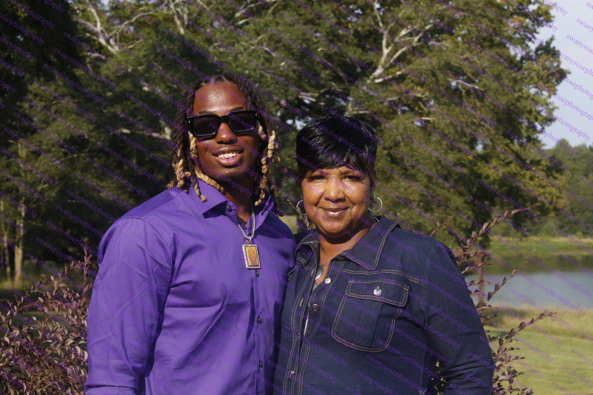 a mother and son standing outdoors in front of trees, smiling. One woman has glasses, long dreadlocks, and is wearing a purple shirt with a necklace. The other woman has short dark hair and is wearing a dark denim jacket.