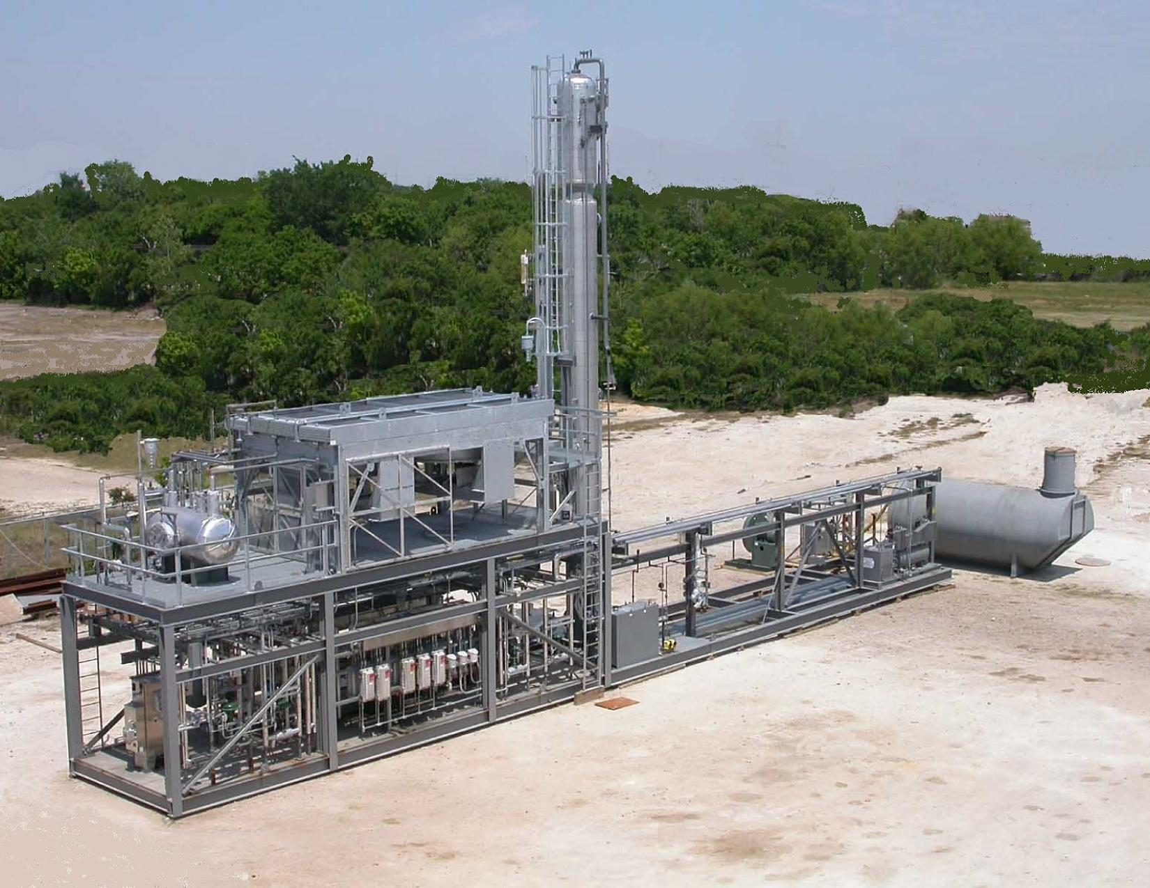 Industrial equipment on a dirt landscape with green trees in the background, including a tall vertical pipe and various metallic structures.
