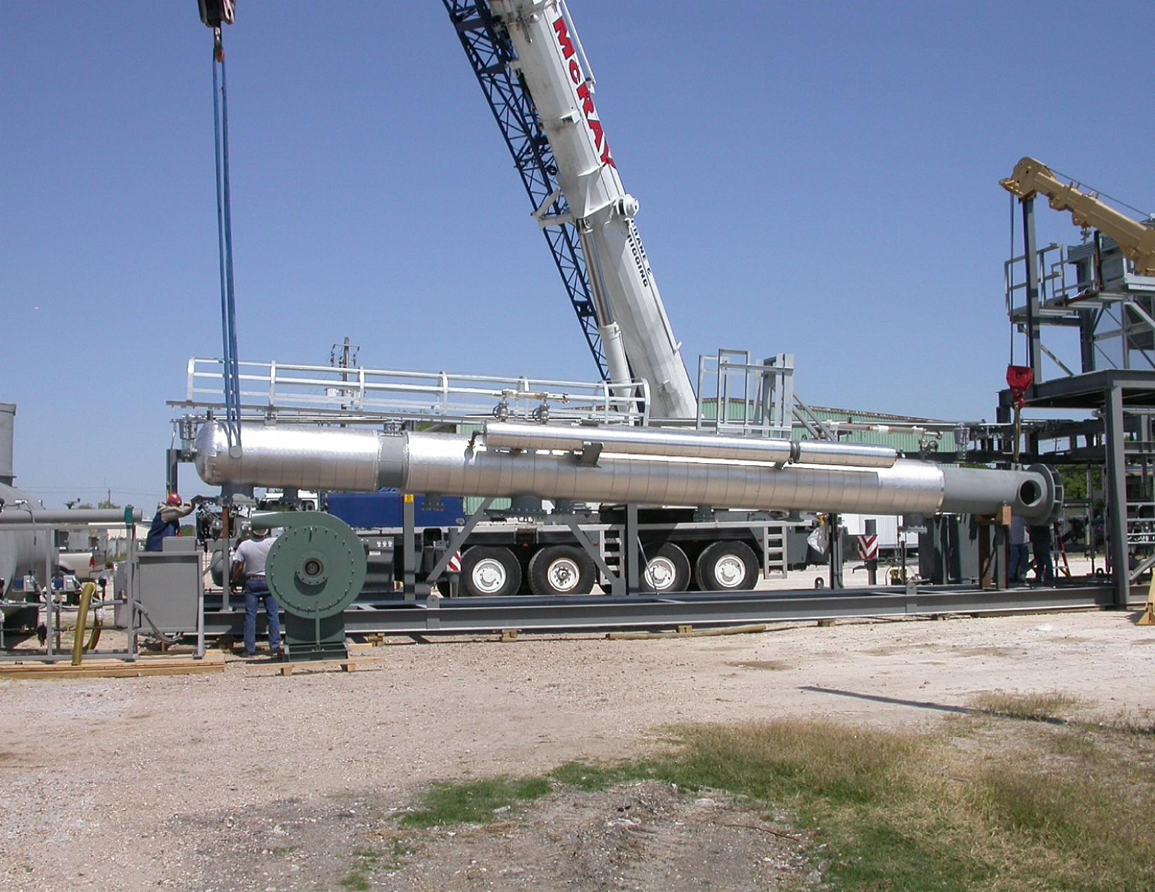 A large industrial pipeline being assembled outdoors with a crane lifting sections into place, and workers nearby supervising the process.