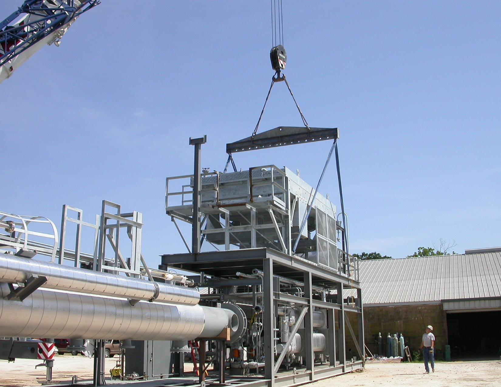 Industrial facility with metal pipes, a crane lifting a large metal structure, and a worker wearing a helmet standing outside.