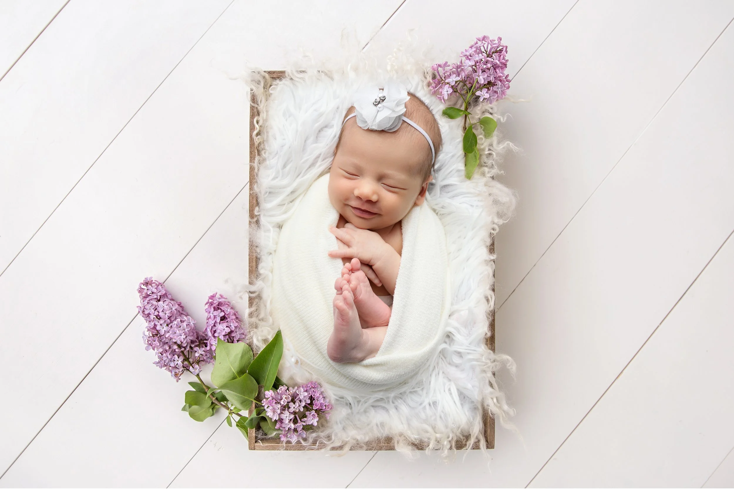A smiling baby lying on a soft white blanket inside a rectangular wooden tray, surrounded by purple lilac flowers on a white wooden floor.