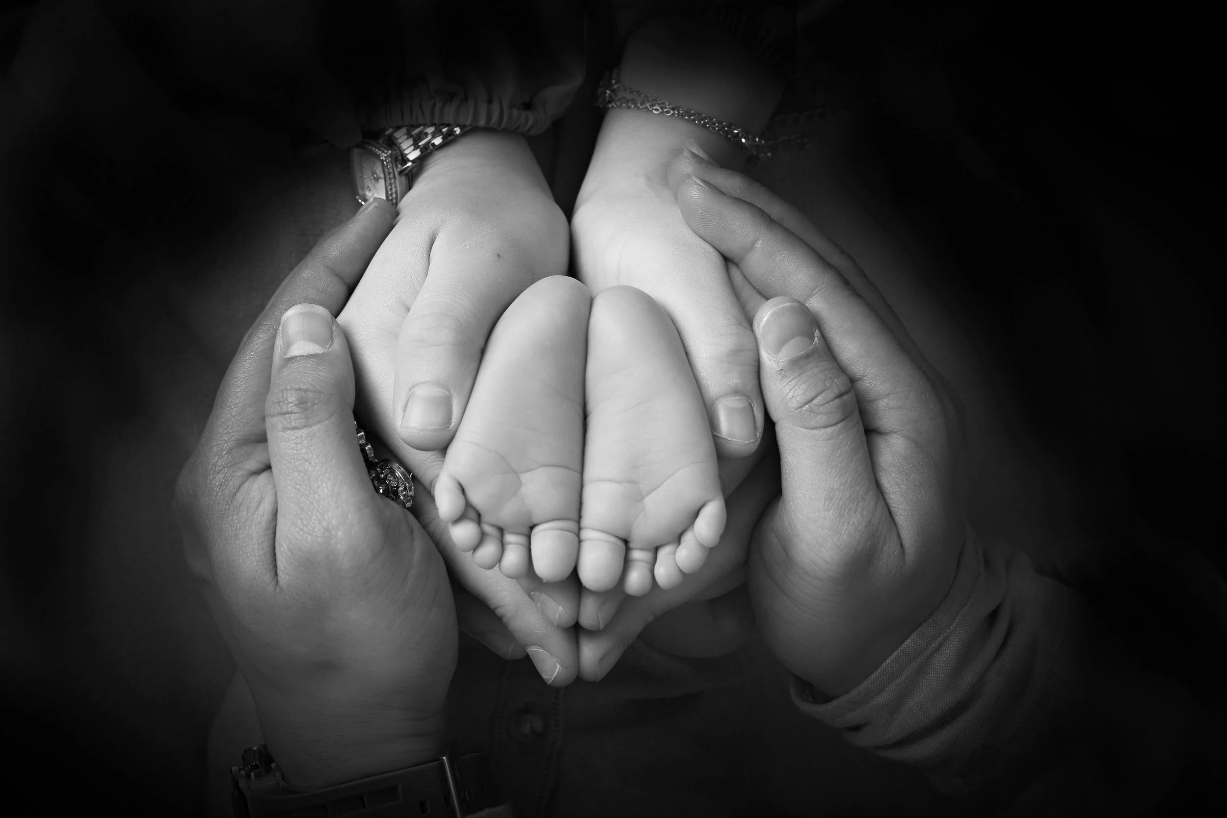 A black and white photo of multiple hands holding a small child's hand, with some hands adorned with watches and bracelets.