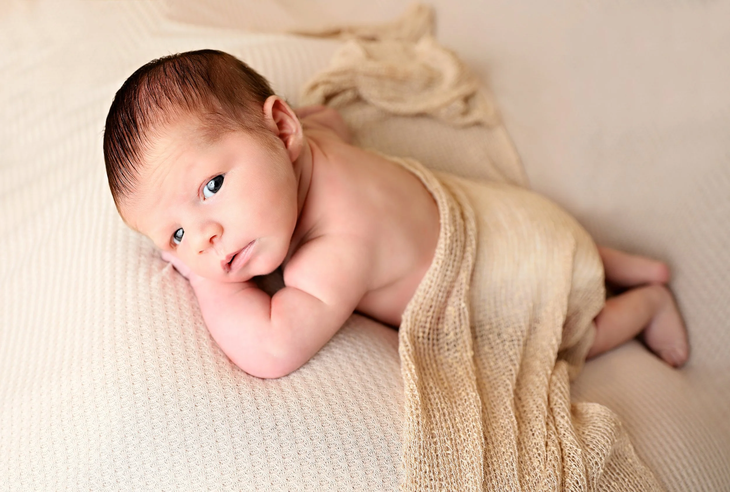 A close-up of a newborn baby lying on a soft, beige blanket, looking slightly to the side with wide eyes and minimal clothing, with one arm bent and hand near the face.