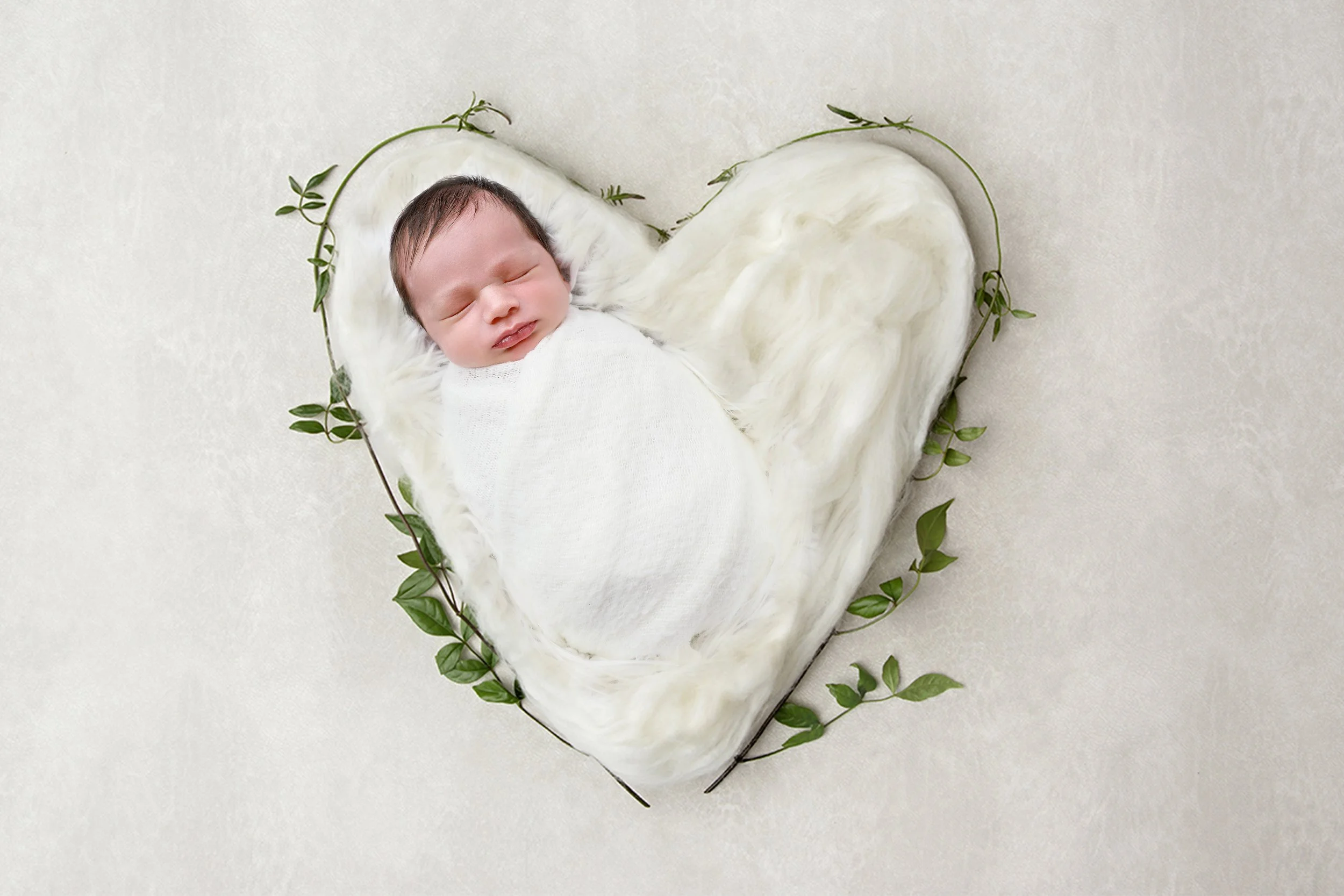A newborn baby with dark hair and light skin, peacefully sleeping on a white furry blanket shaped like a heart, with a green leafy vine surrounding the blanket on a light background.
