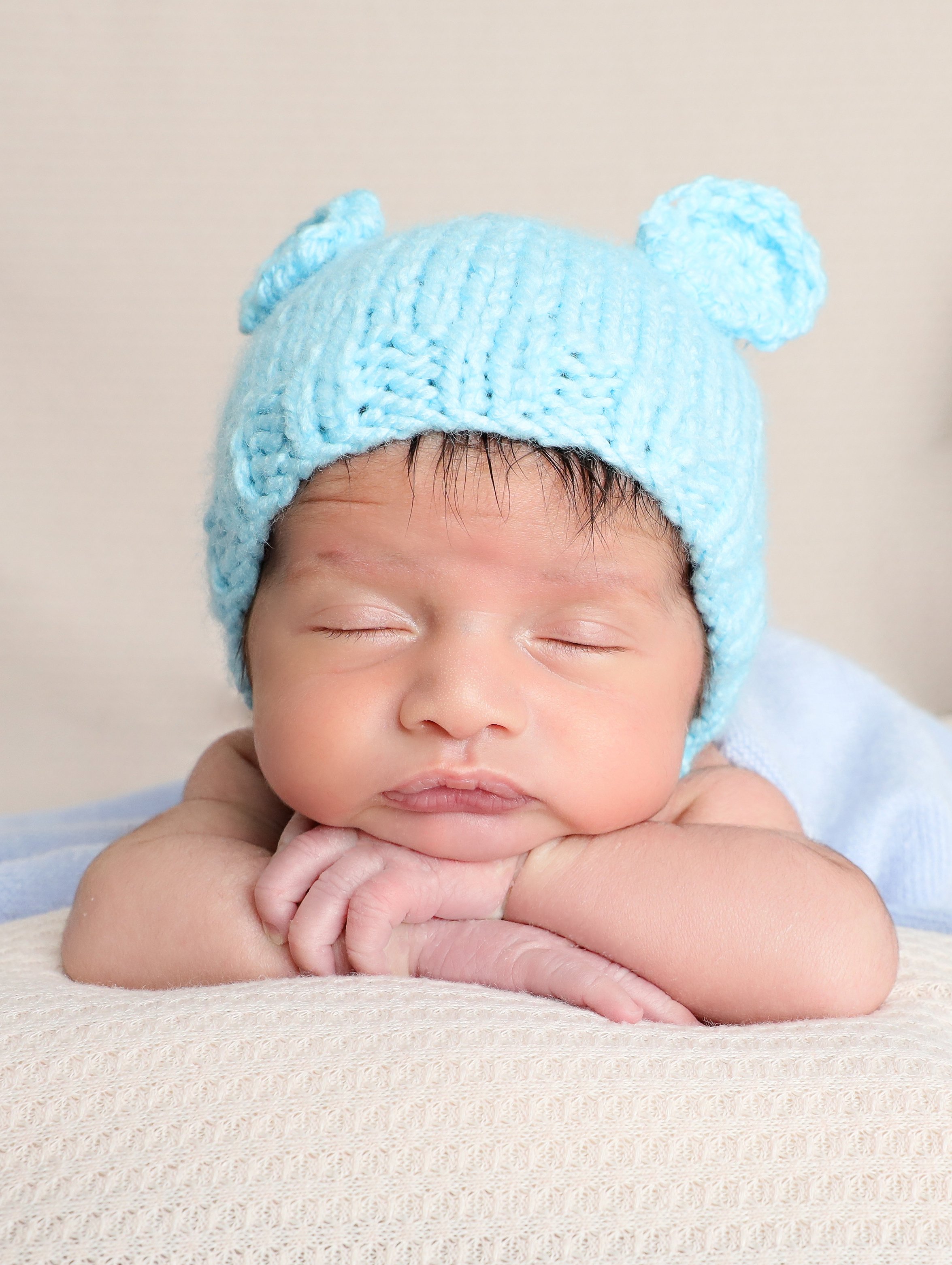 A baby with closed eyes resting on their folded arms, wearing a light blue knit hat with bear ears. The baby has a serene expression and is on a textured beige surface with a plain background.