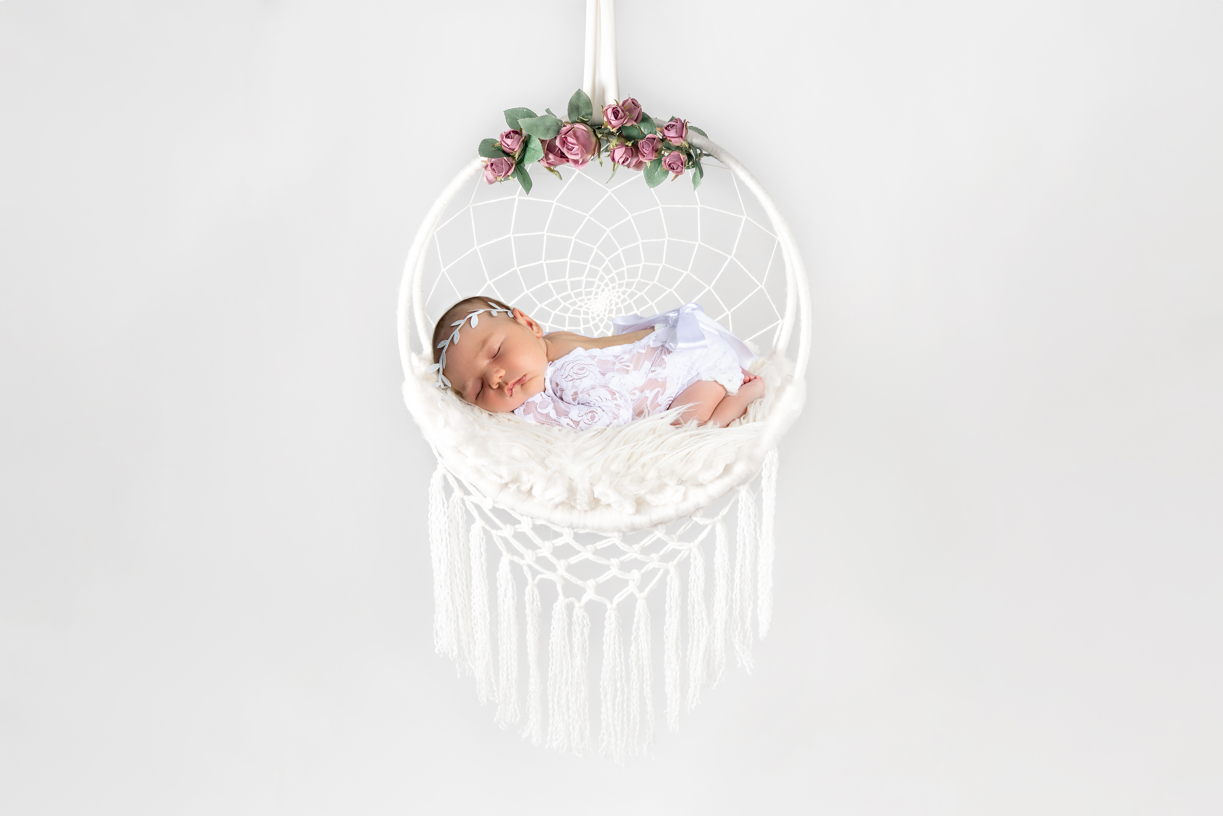 A sleeping baby girl lying in a white hammock with pink roses and green leaves decoration, against a plain white background.