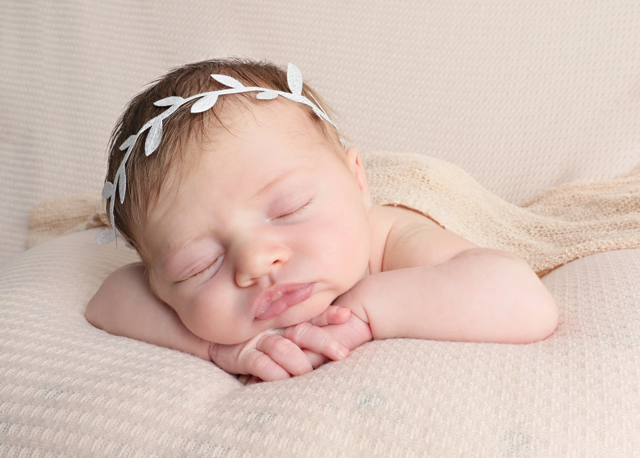 A sleeping baby with a silver leaf-shaped headband resting on a soft beige surface.