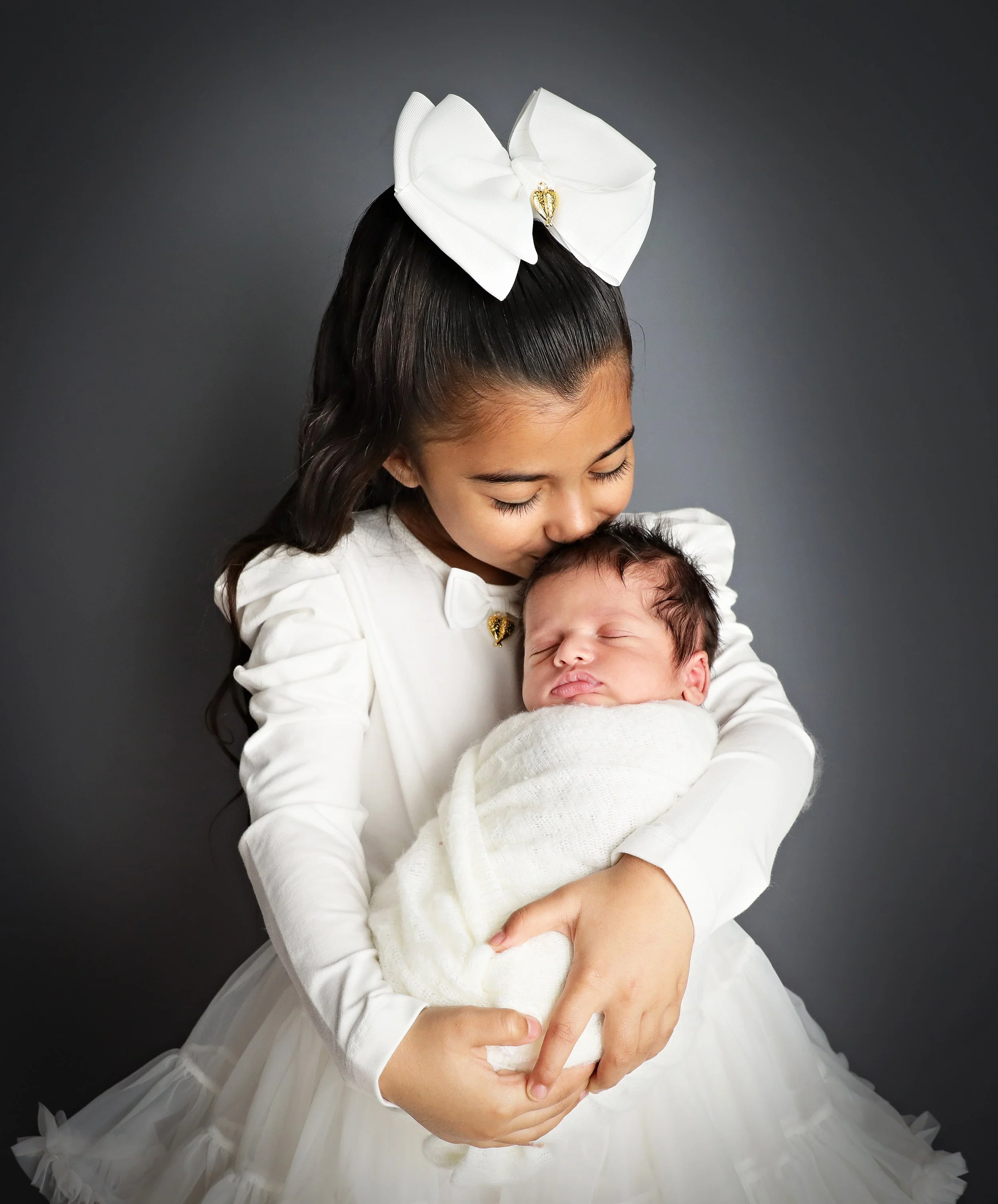 A young girl with long dark hair, dressed in a white dress with a large white bow on her head, holding a newborn baby wrapped in a white blanket, both against a dark gray background.
