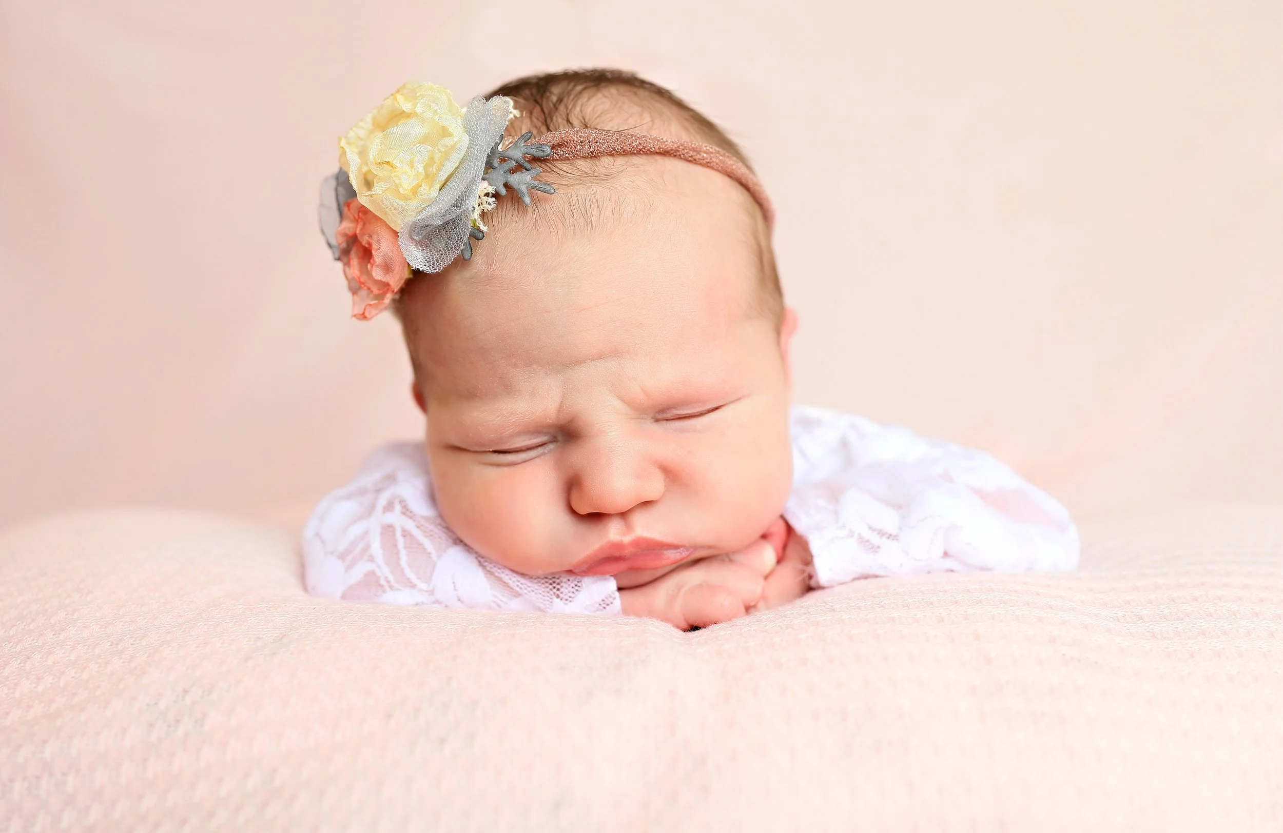 Close-up of a sleeping baby with a floral headband and a white lacy outfit, resting on a soft pink surface.